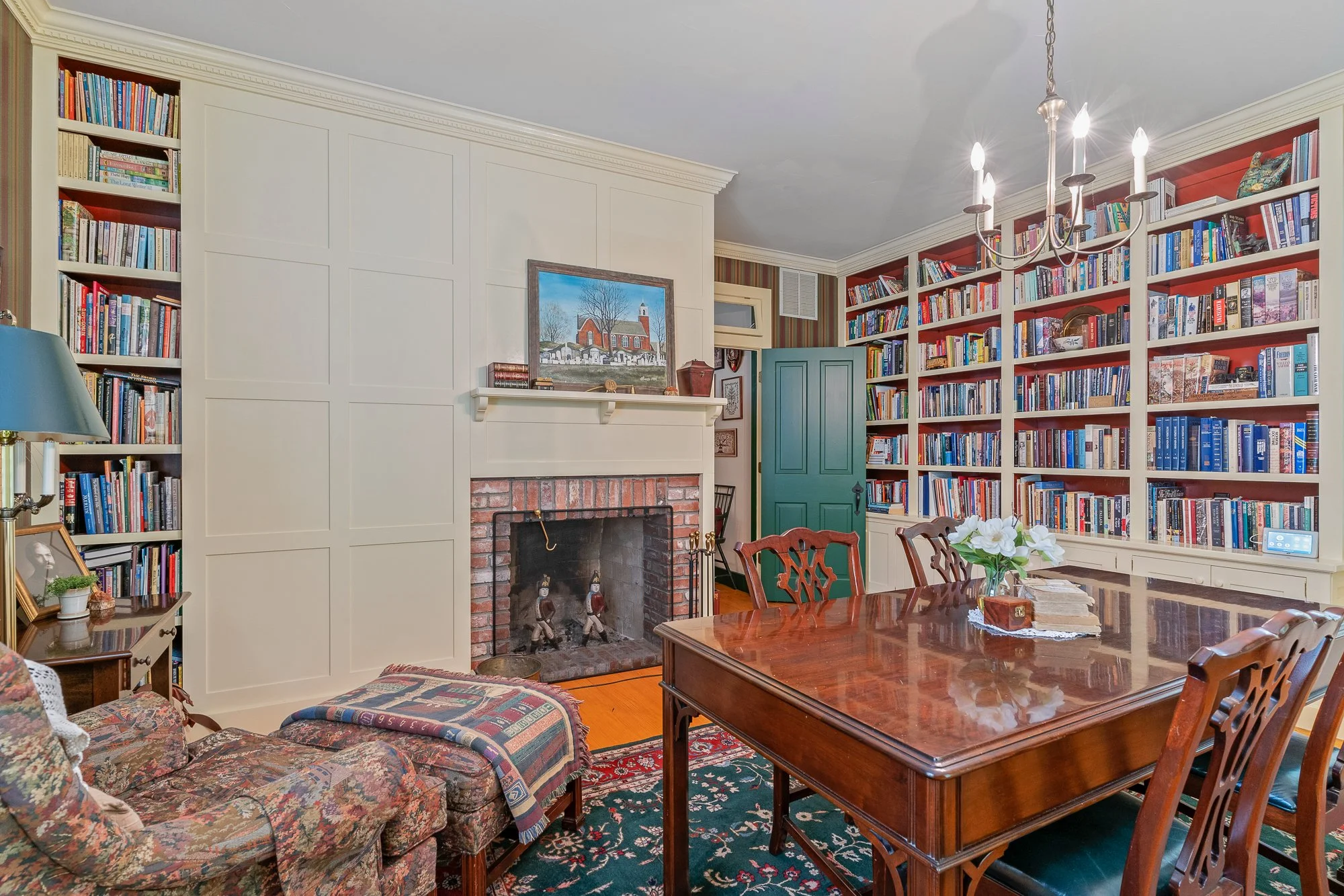 Living room with a brick fireplace, white built-in bookshelves filled with books, a wooden dining table with chairs, a chandelier, and various decorative items.