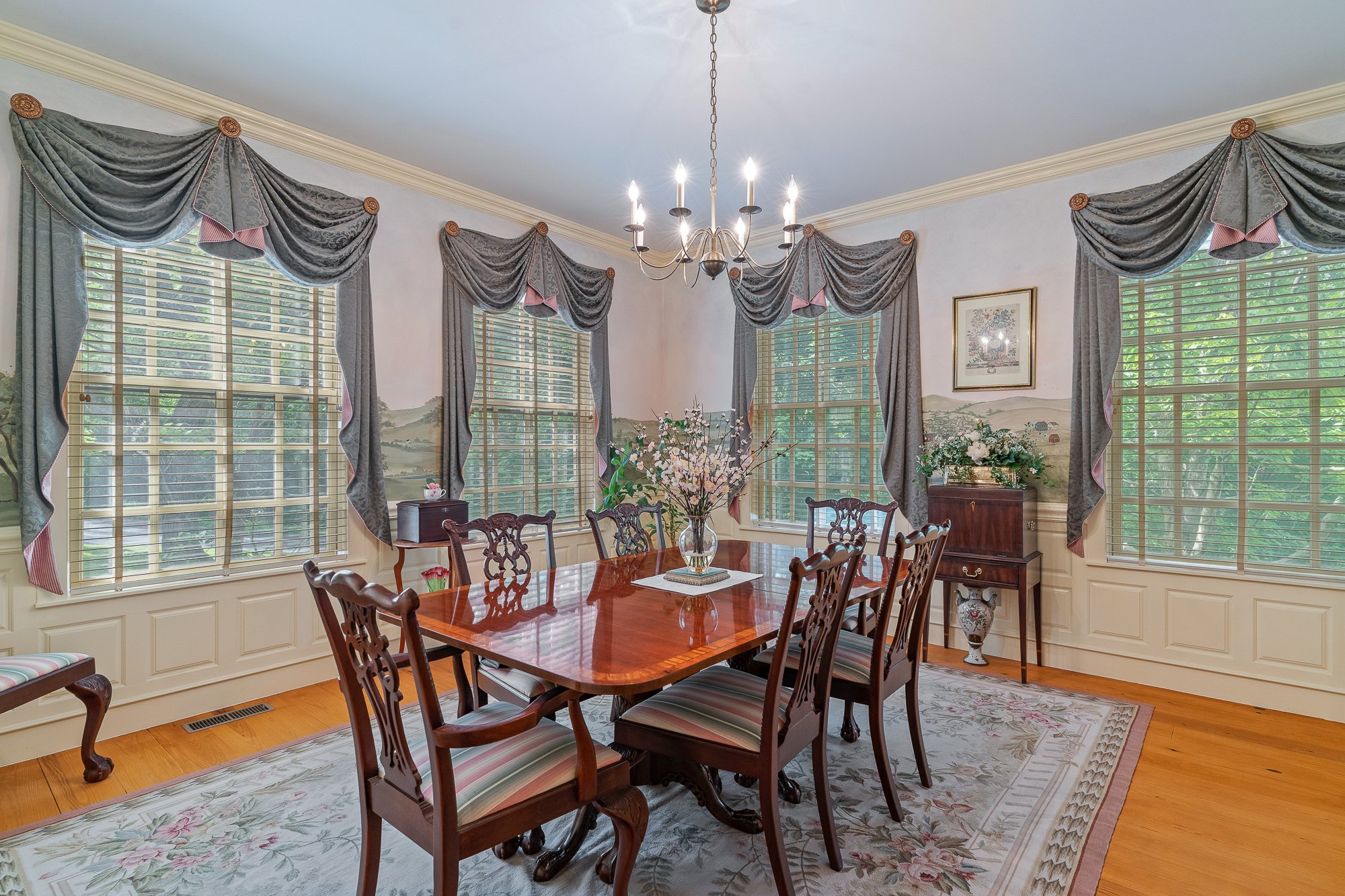 A traditional dining room with large windows and elegant drapes, a wooden dining table with chairs, a chandelier overhead, and floral decorations.