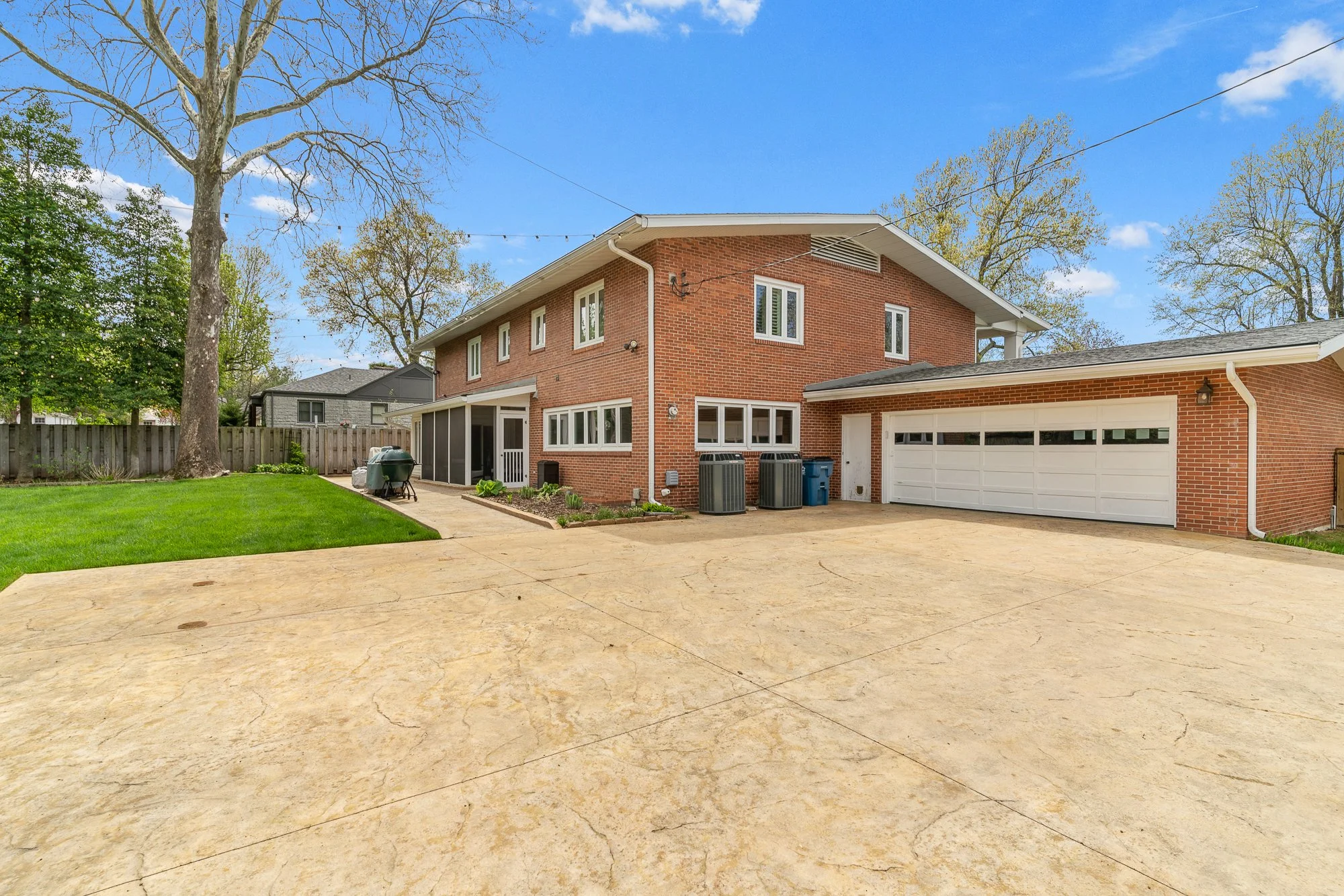 View of a backyard with a large tree, green grass, and a brick house with white trim. The house has a screened porch and a two-car garage. There is a concrete driveway and a fence in the background, with some outdoor furniture and trash bins visible.