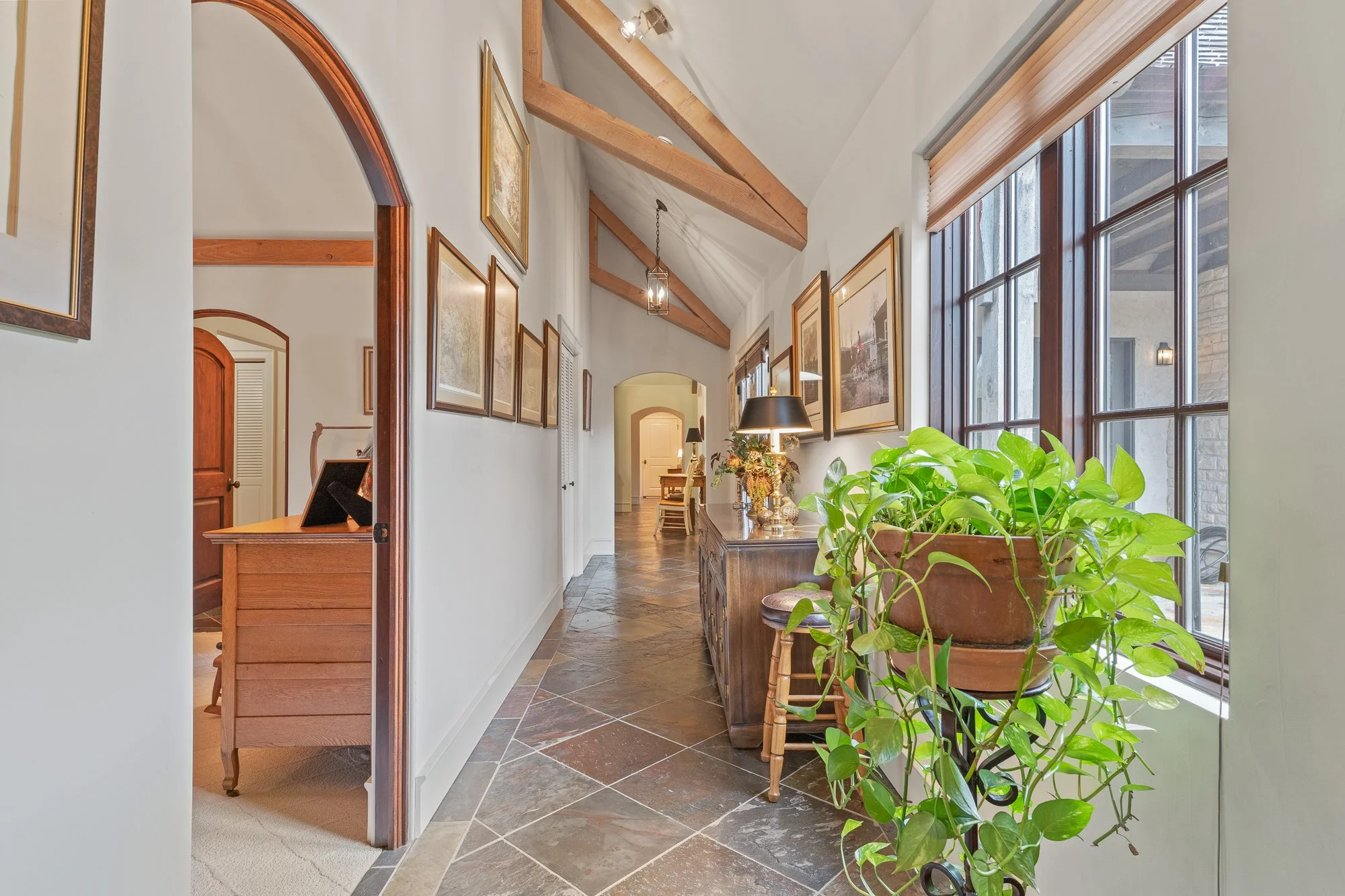 Interior of a house hallway with large windows, wooden beams on ceiling, framed pictures on wall, and a wooden sideboard with plants and lamps.