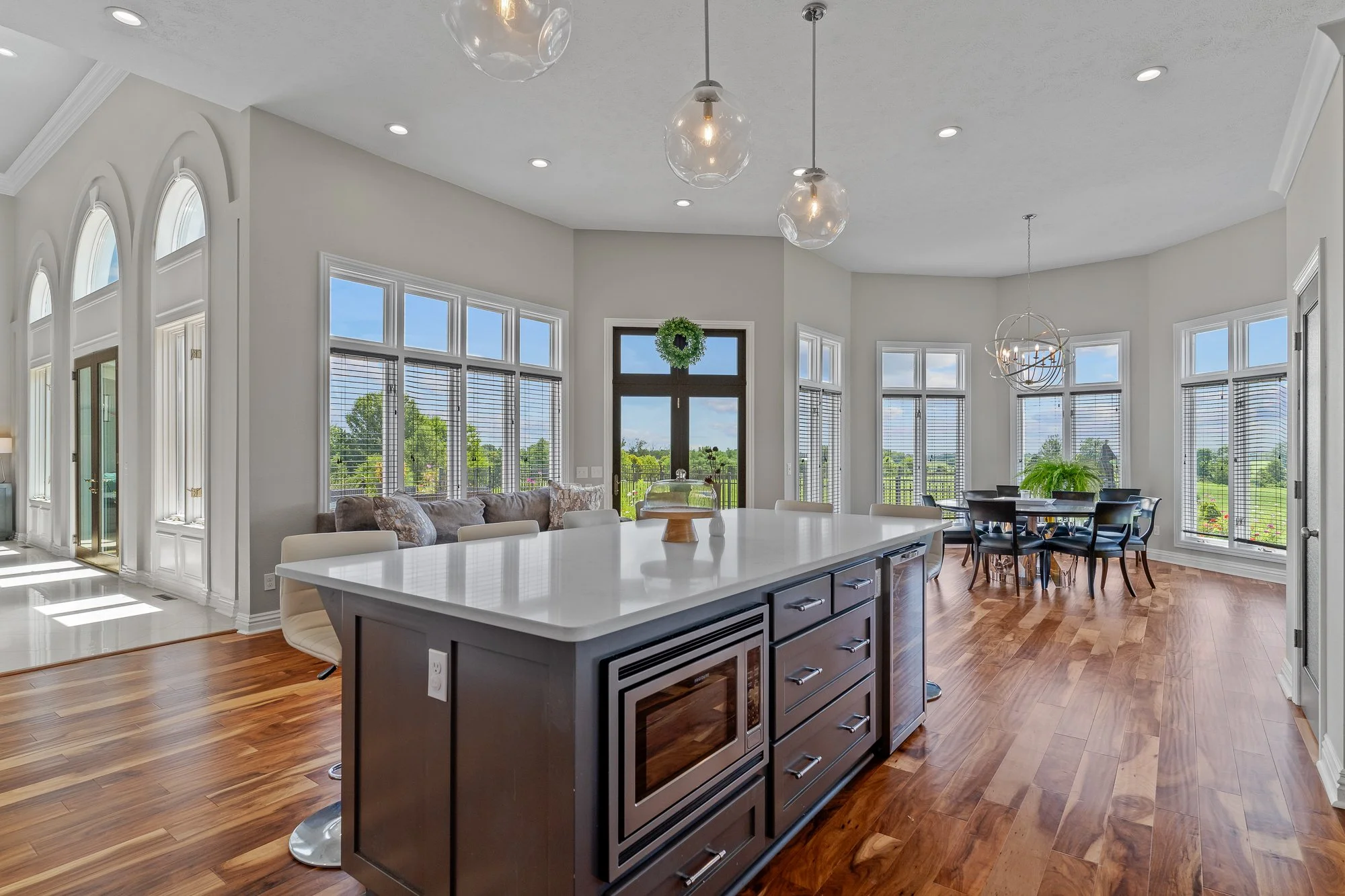 Bright open-concept kitchen and dining area with large windows, wooden floors, a central island with a microwave, living space with a couch, and a green wreath hanging on a door, with views of the outdoors.