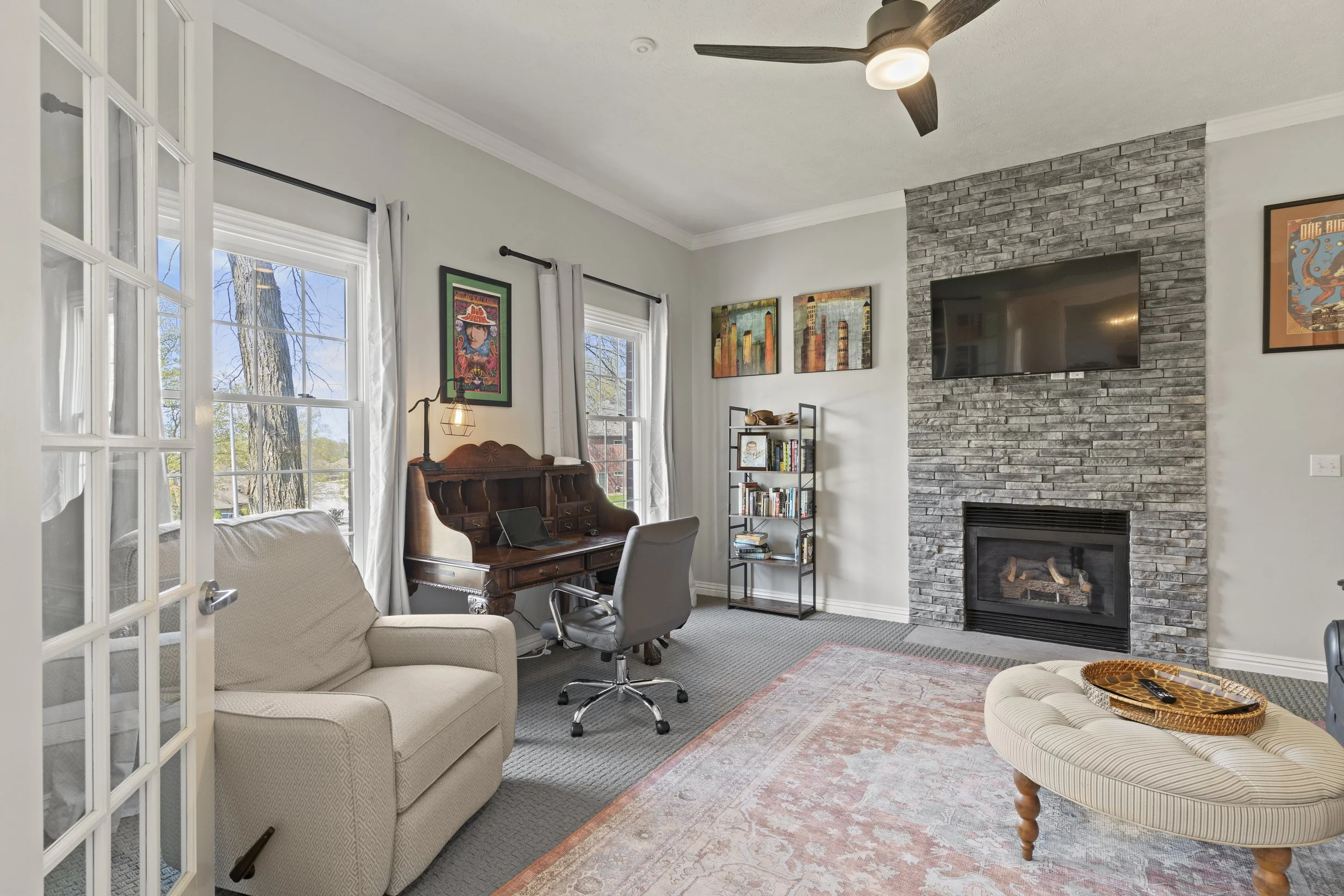 Living room with a cream sofa, a patterned armchair with a tray, two windows with white curtains, framed artwork on the wall, a wooden desk with lamp and a gray office chair, a black metal bookshelf, a gray brick fireplace with a TV mounted above, wo