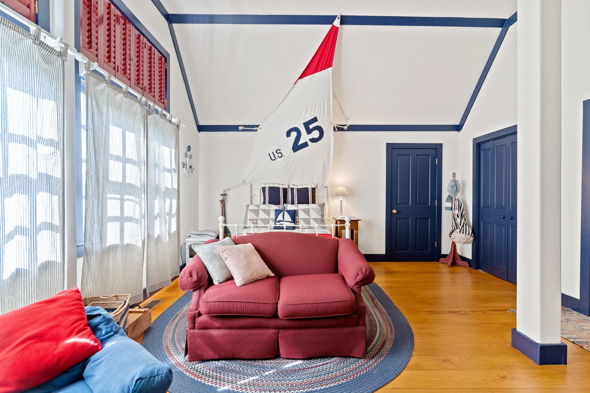 A bedroom with white walls and blue trim, a bed with red, white, and blue bedding, a small wooden nightstand with a lamp, a white bed with a large U.S. Navy sailboat flag as a headboard, a red couch with striped pillows, windows with white and gray s