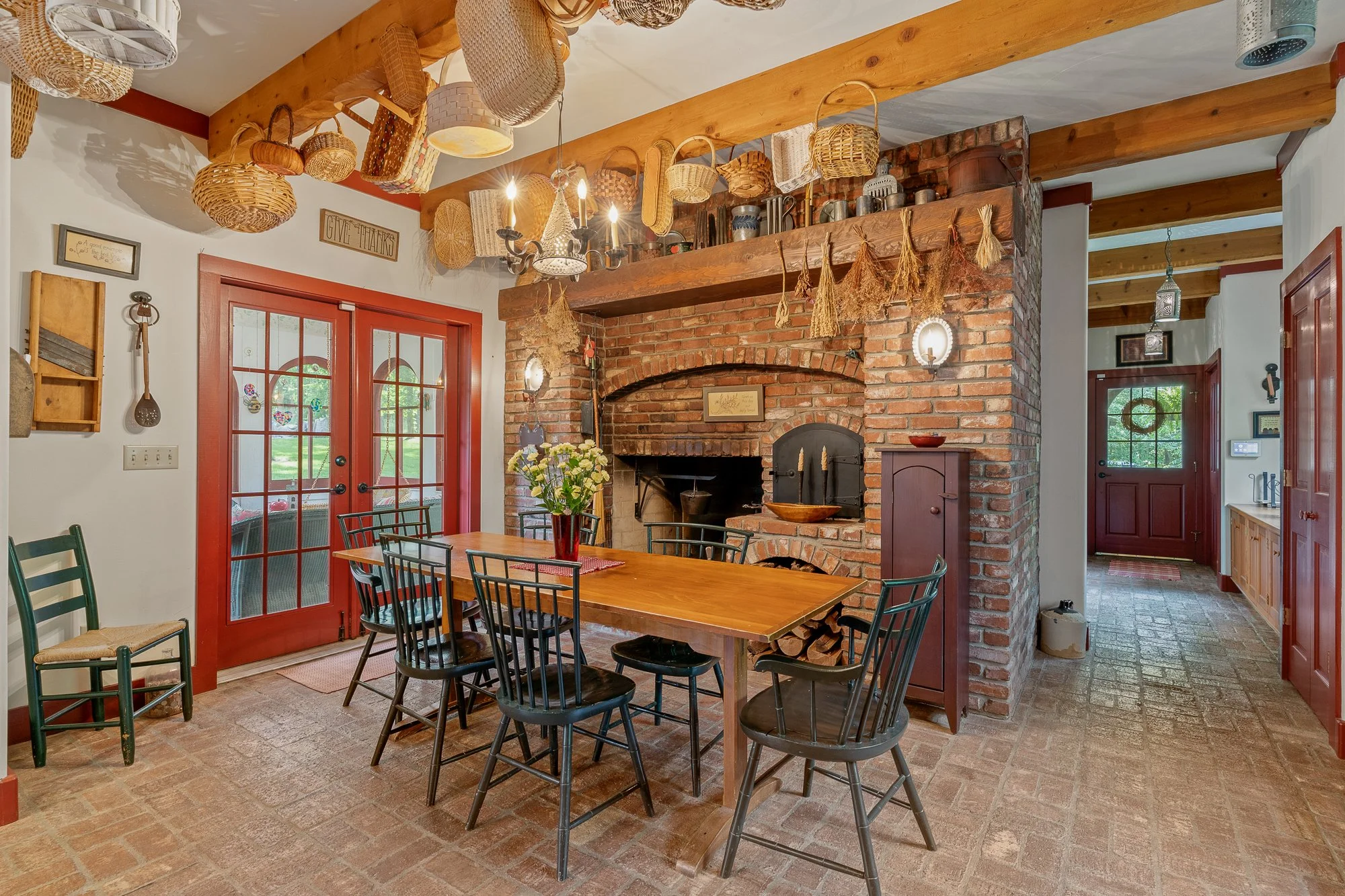 A cozy kitchen and dining area with a brick fireplace, a wooden dining table surrounded by black chairs, and hanging baskets and decorations on the ceiling. There are red double doors with glass panes leading outside, and a hallway with brick floorin