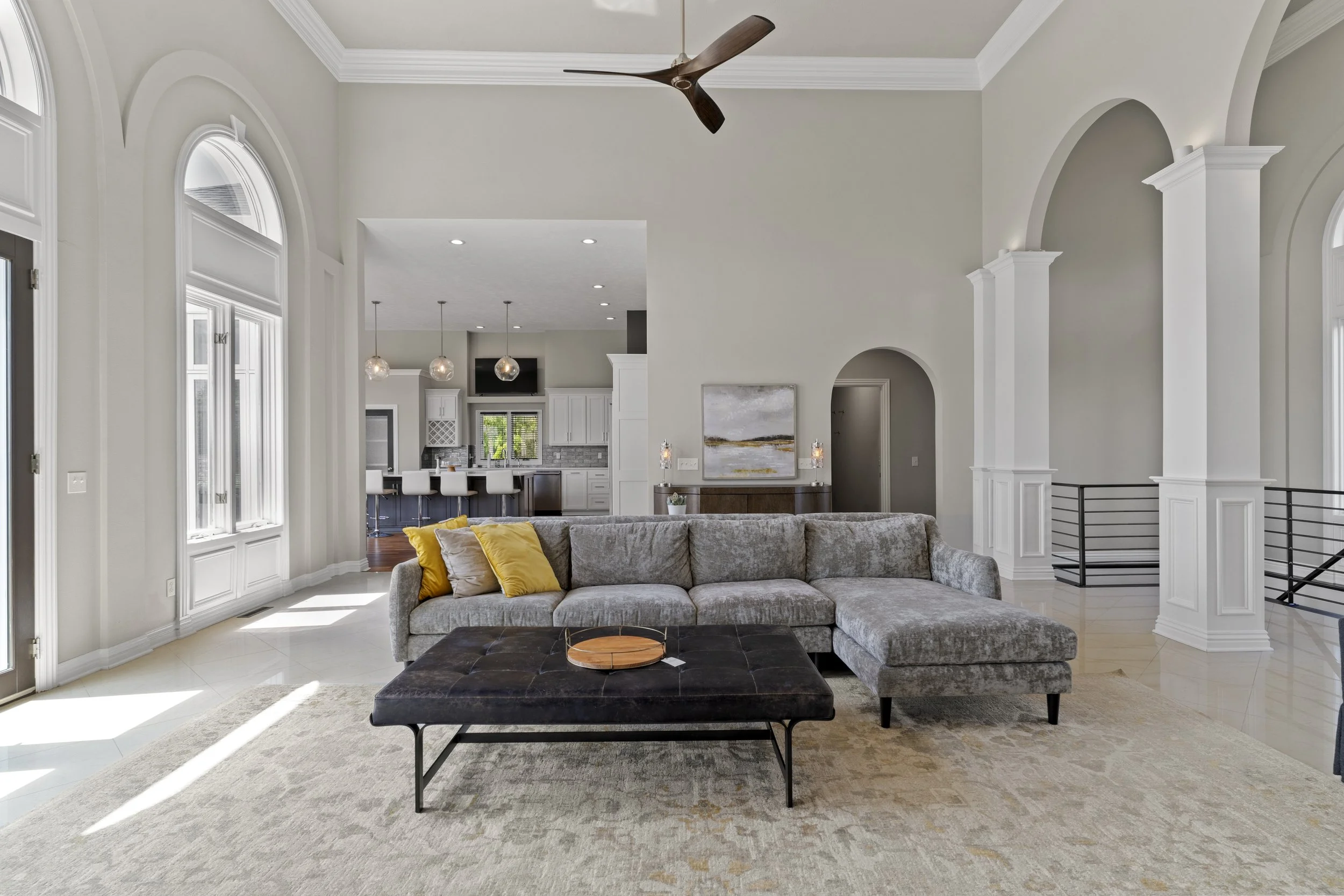 Spacious living room with large windows, a gray sectional sofa with yellow and gray pillows, black ottoman, light-colored rug, open kitchen with bar stools, pendant lights, flat-screen TV, and artwork.