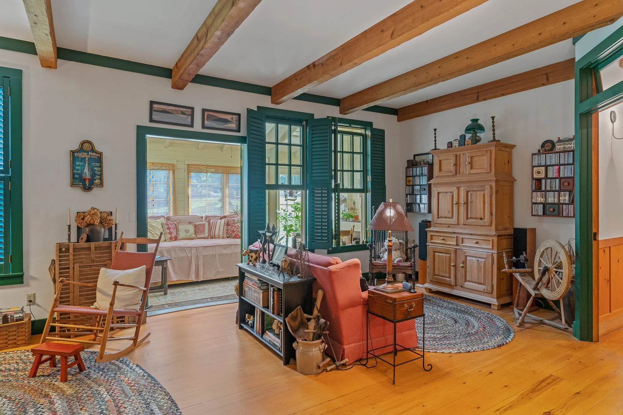 Living room with wooden ceiling beams, green window shutters, a wooden cabinet, and various furniture including a red armchair, a small wooden chair, and a coffee table with decorative items. There are two framed artworks and a CD rack on the wall, a