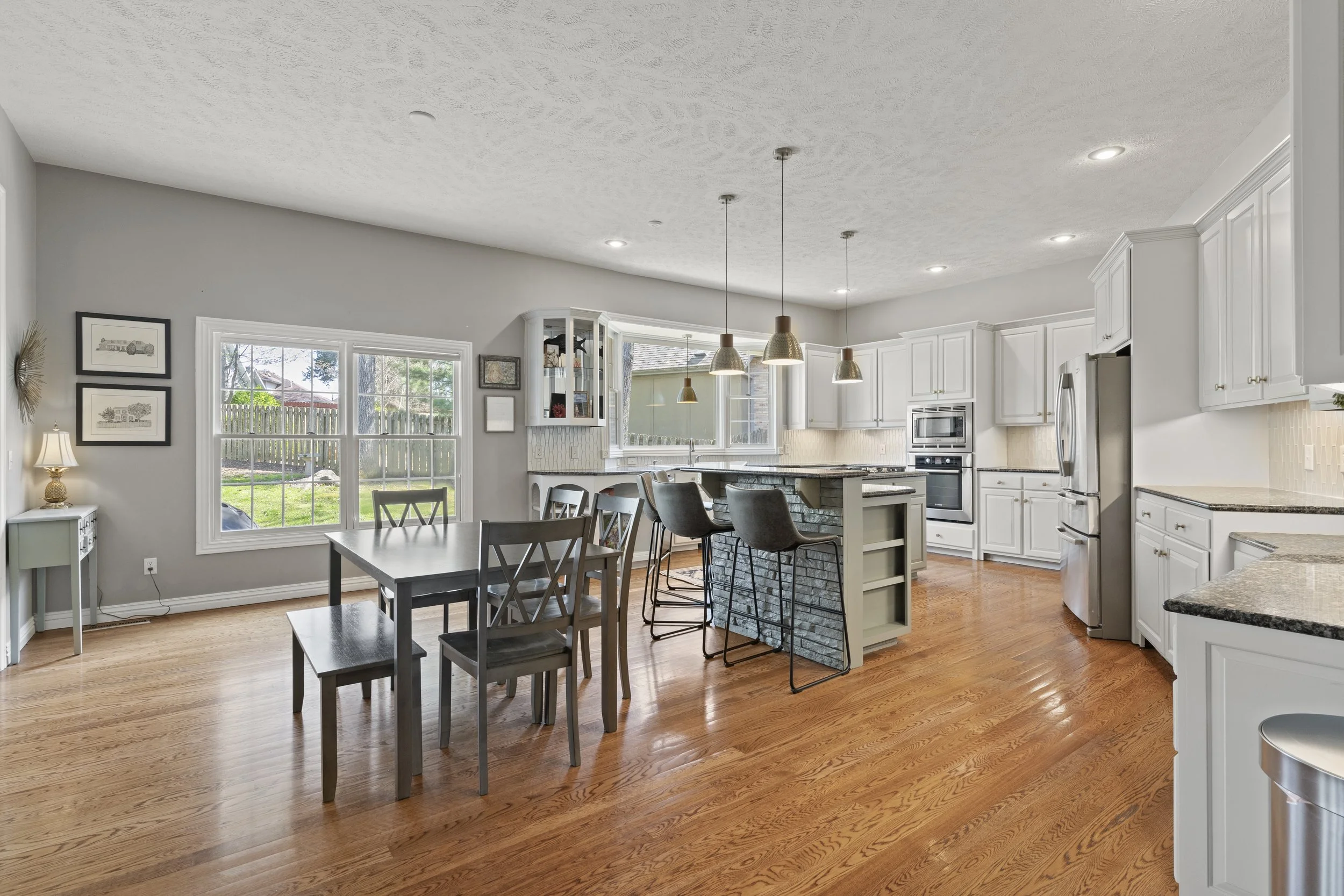 A spacious open-concept kitchen and dining area with wooden floors, white cabinets, stainless steel appliances, a kitchen island with bar stools, a dining table with chairs, large windows showing a backyard, and pendant lights hanging above the islan
