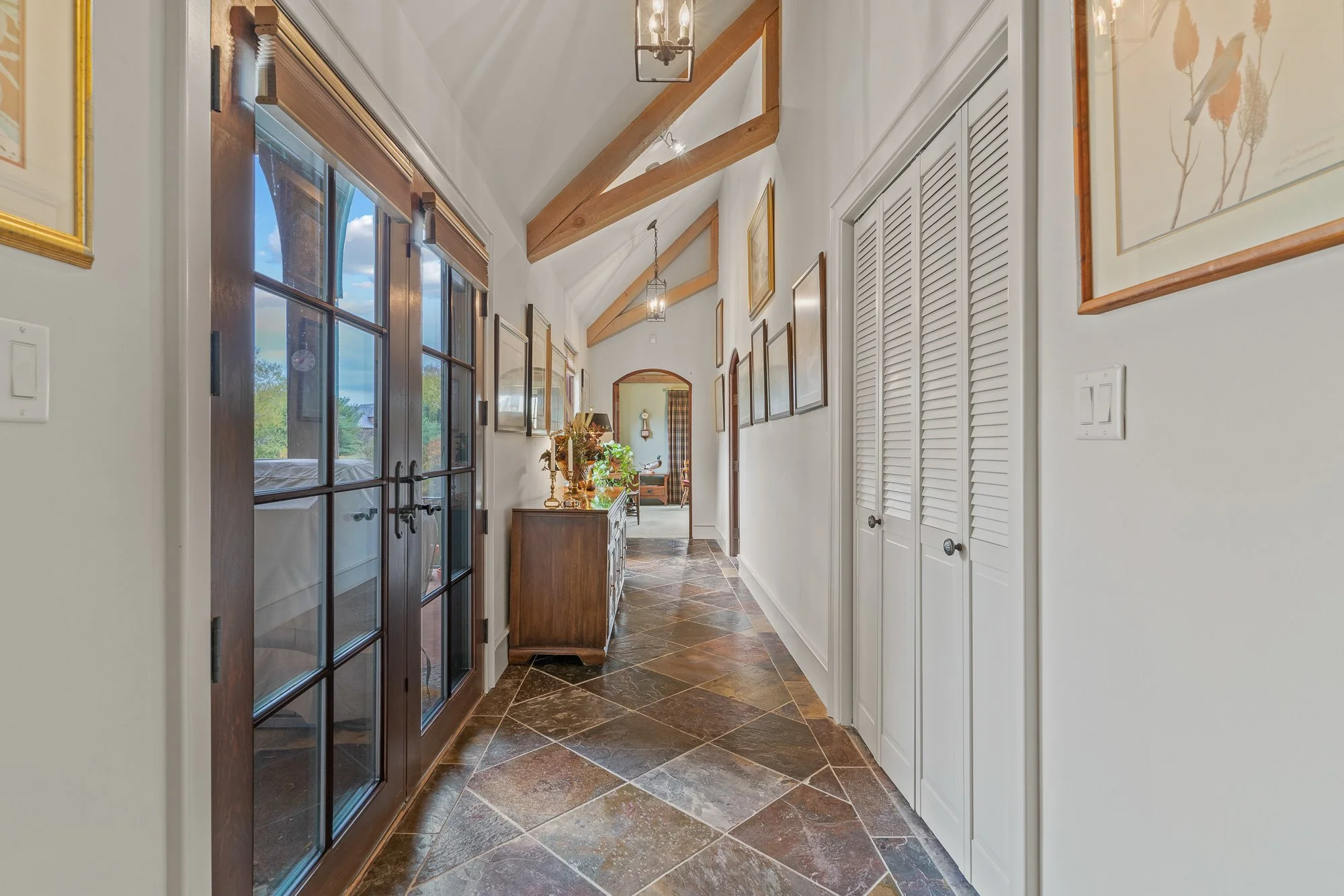 A hallway with a wooden front door featuring glass panes, beige walls decorated with framed artwork, wooden beams on the ceiling, and a tiled floor. Sunlight is coming through the windows.