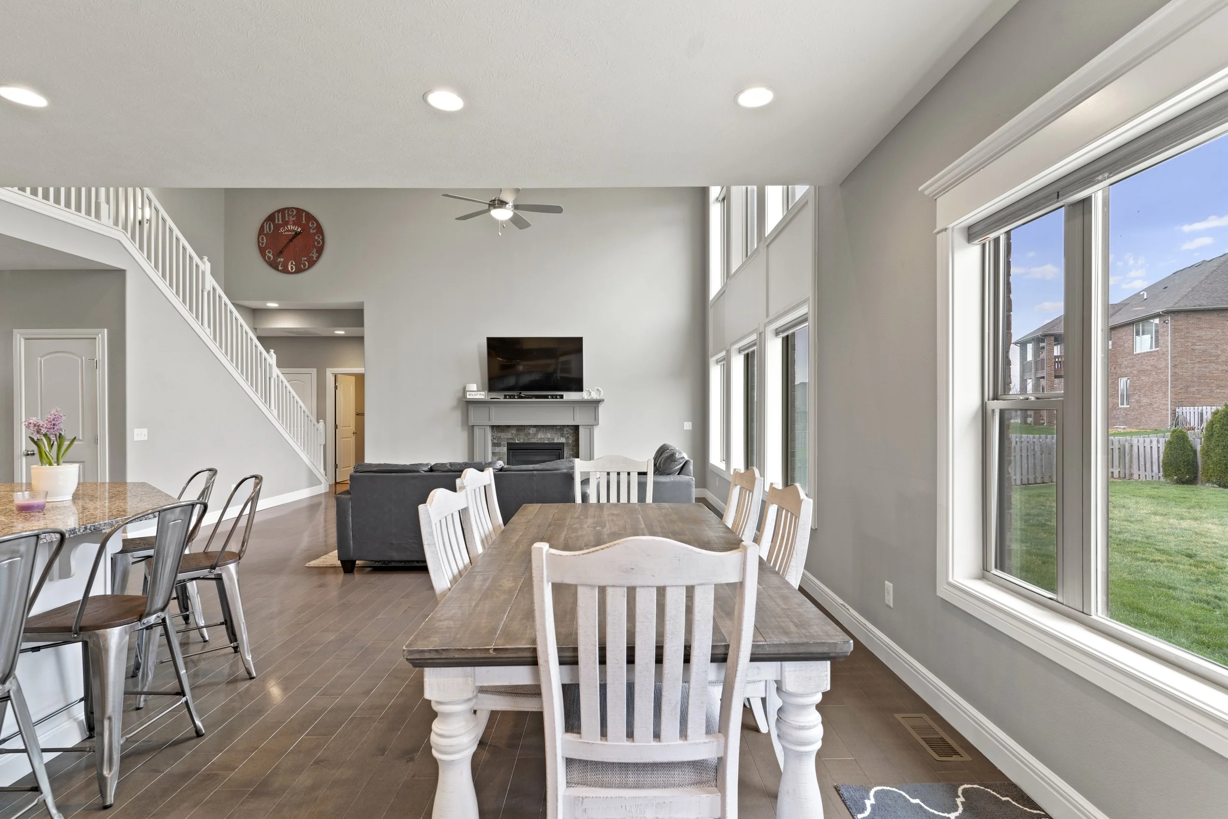 Open-concept living and dining area with large windows, a fireplace, mounted flat-screen TV, dining table with white chairs, and kitchen counter with metal bar stools.