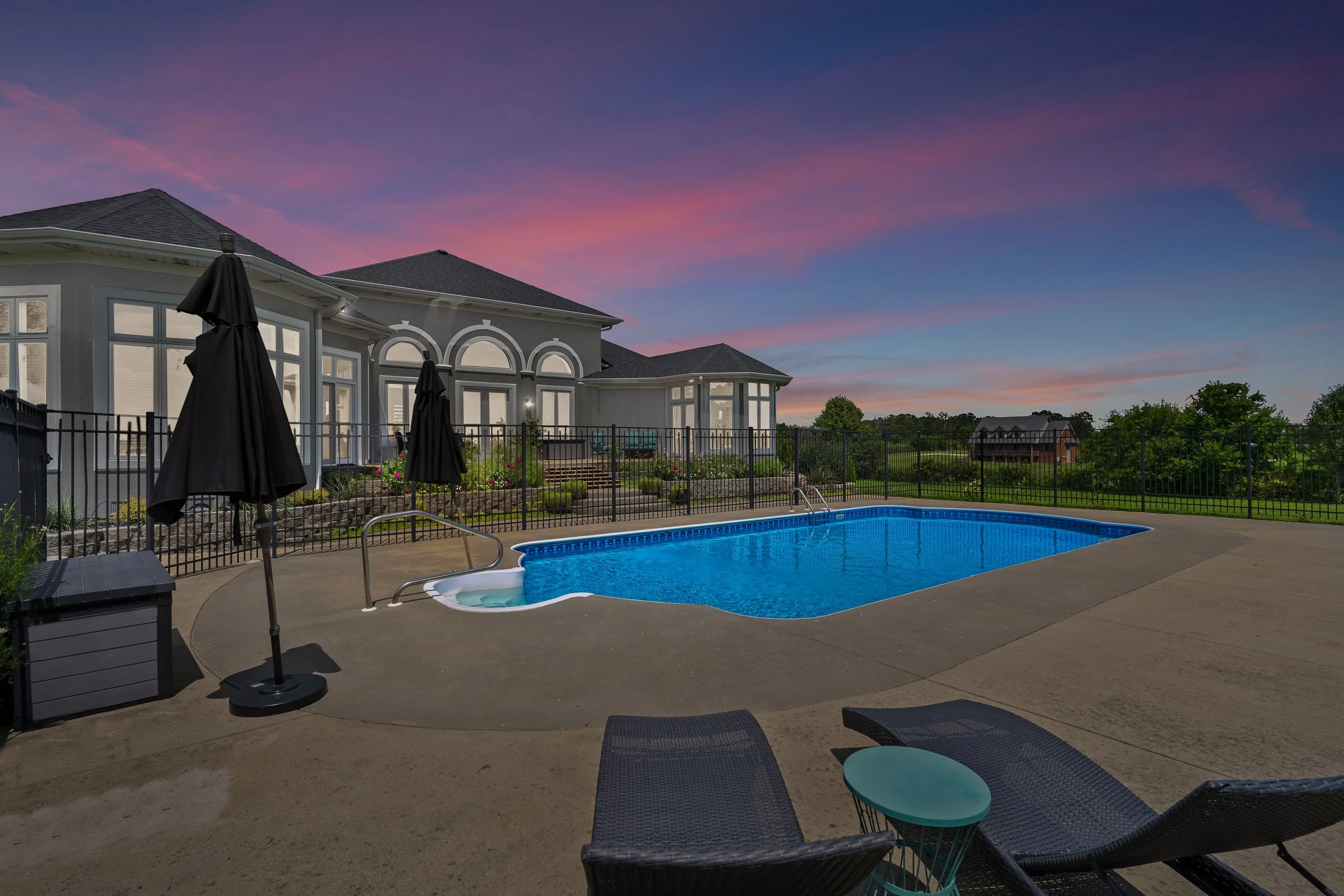 A backyard view of a house with large windows and a swimming pool during dusk, with black poolside umbrellas, lawn furniture, and a landscaped garden, under a sky with pink and purple clouds.