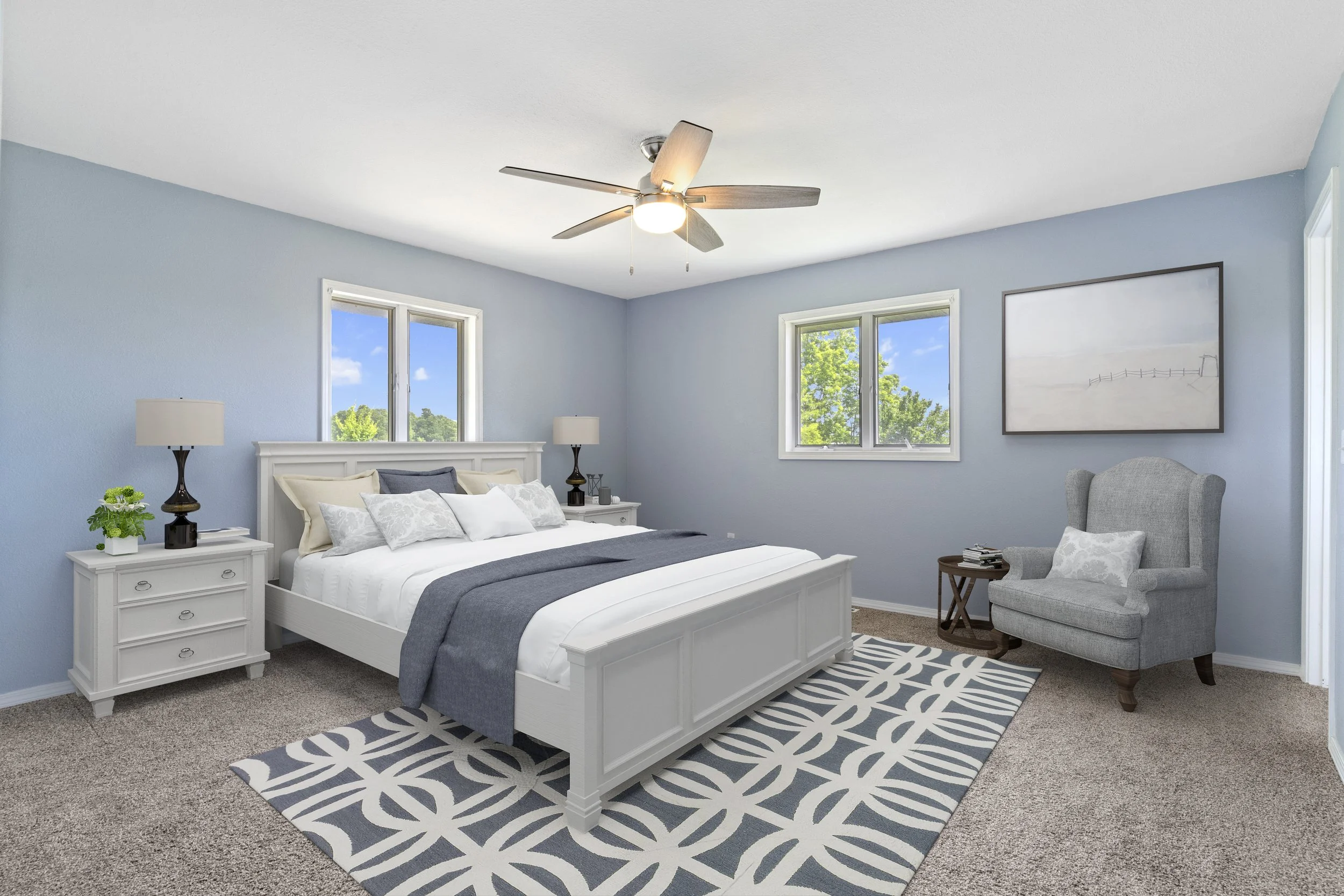A bedroom with light blue walls, a white bed with blue and white bedding, two nightstands with lamps, a gray armchair, a large window showing green trees and blue sky, a framed artwork on the wall, and a patterned area rug.
