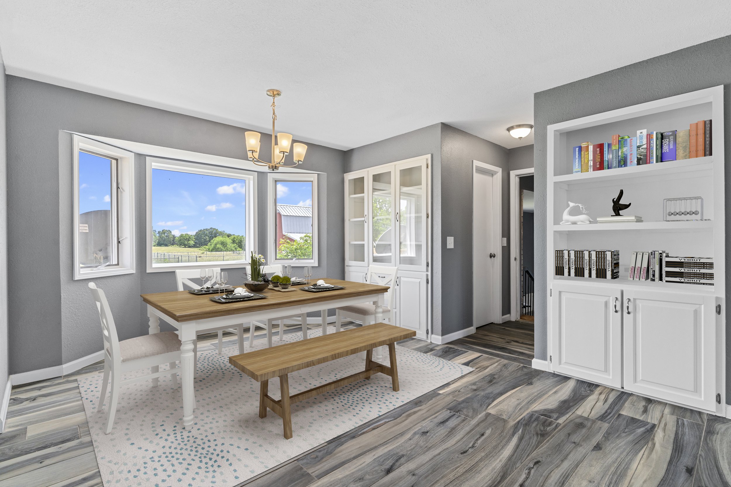 A bright dining area featuring a wooden table set with four place settings with glassware, plates, and napkins, surrounded by white chairs and a bench. There is a large window with a scenic view of greenery and a building, and a white china cabinet w