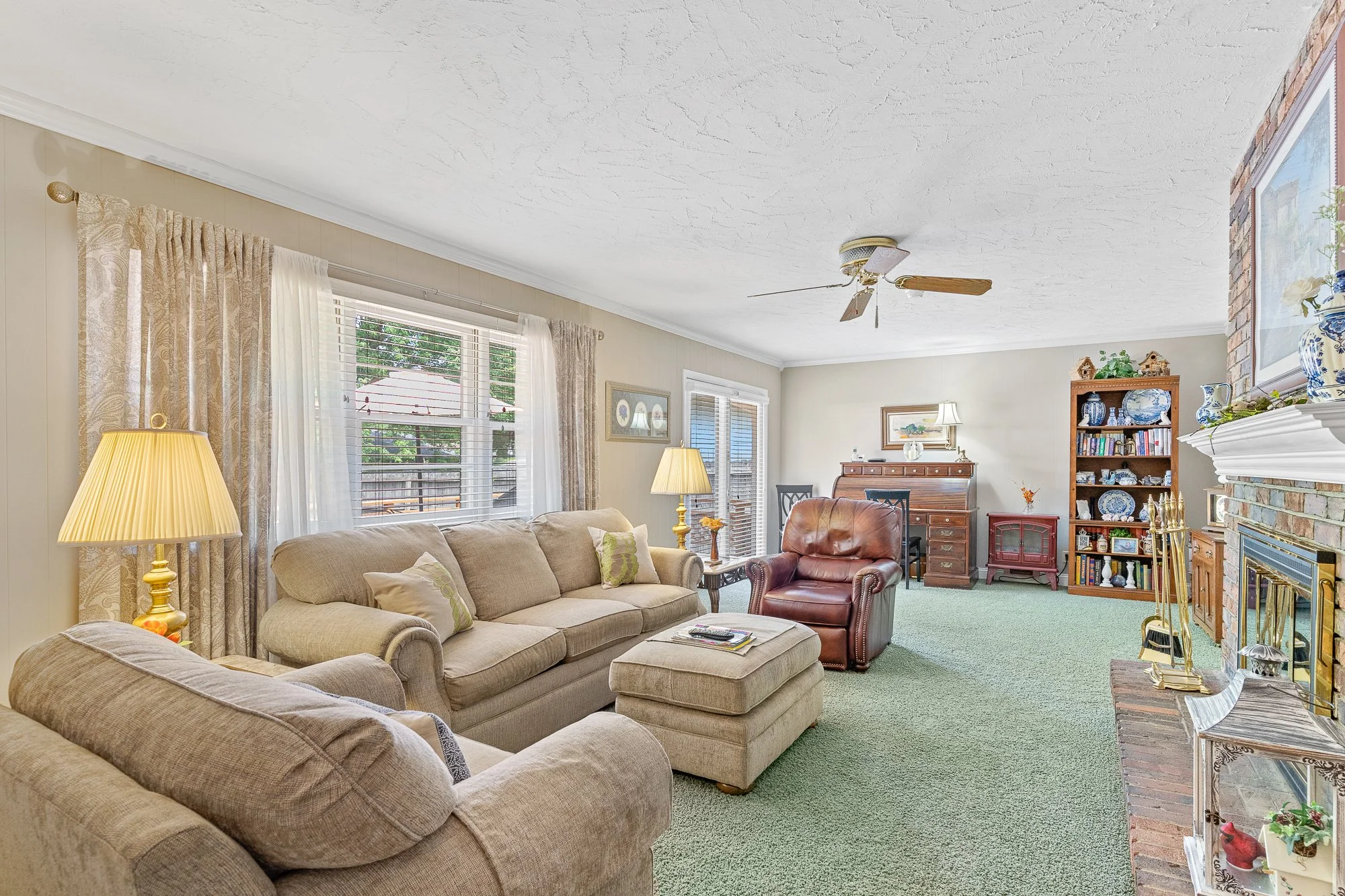 A living room with beige sofa and pillows, a red armchair, and a green carpet. There are windows with curtains, a wooden bookshelf with decorative items and books, a ceiling fan, a brick fireplace with a white mantel, and various lamps and decoration