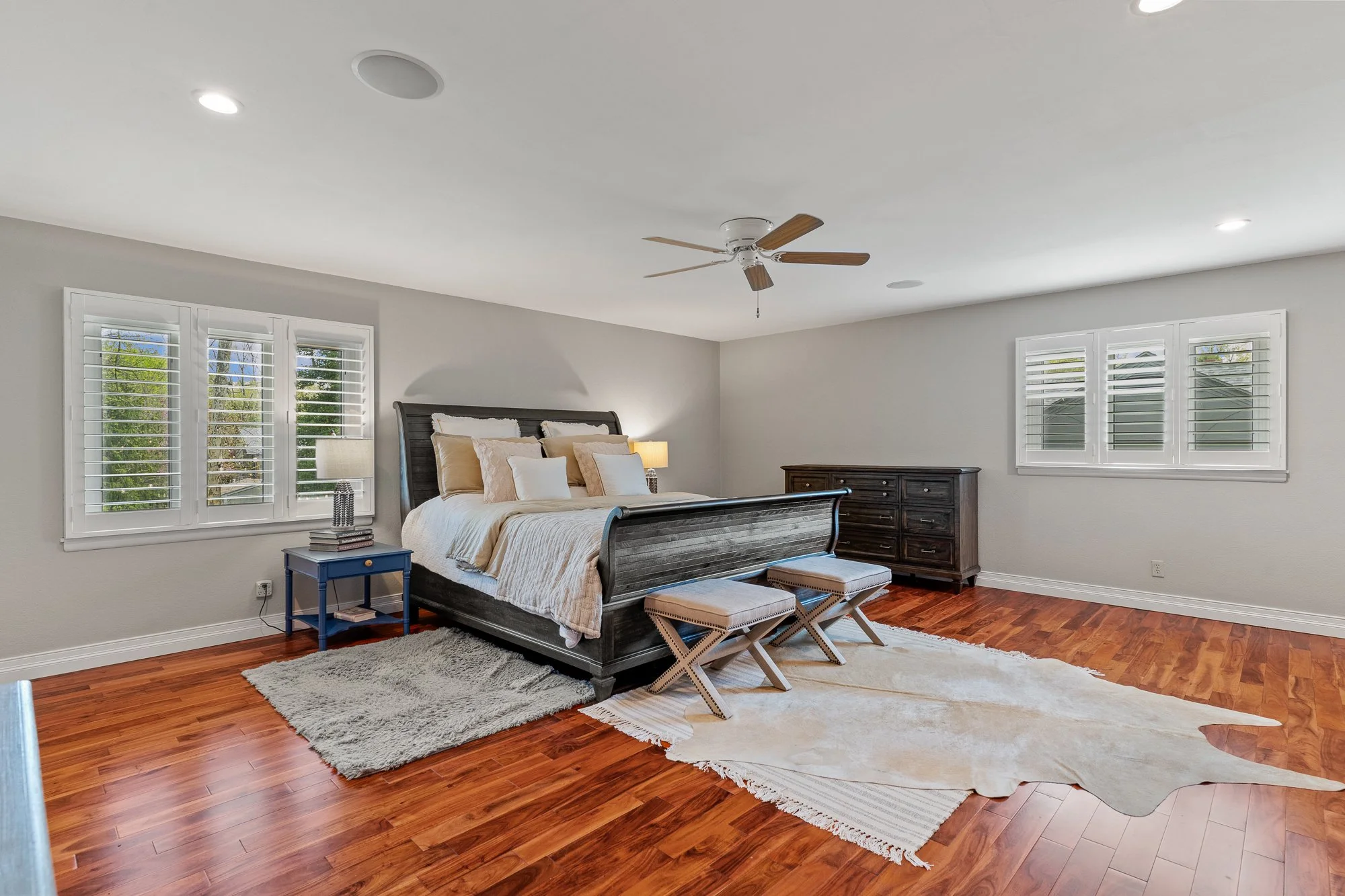 A bedroom with a dark wood bed frame, white and beige bedding, two matching ottomans at the foot, a blue nightstand with books, a beige rug, and multiple windows with white shutters, wooden floors, and a ceiling fan.