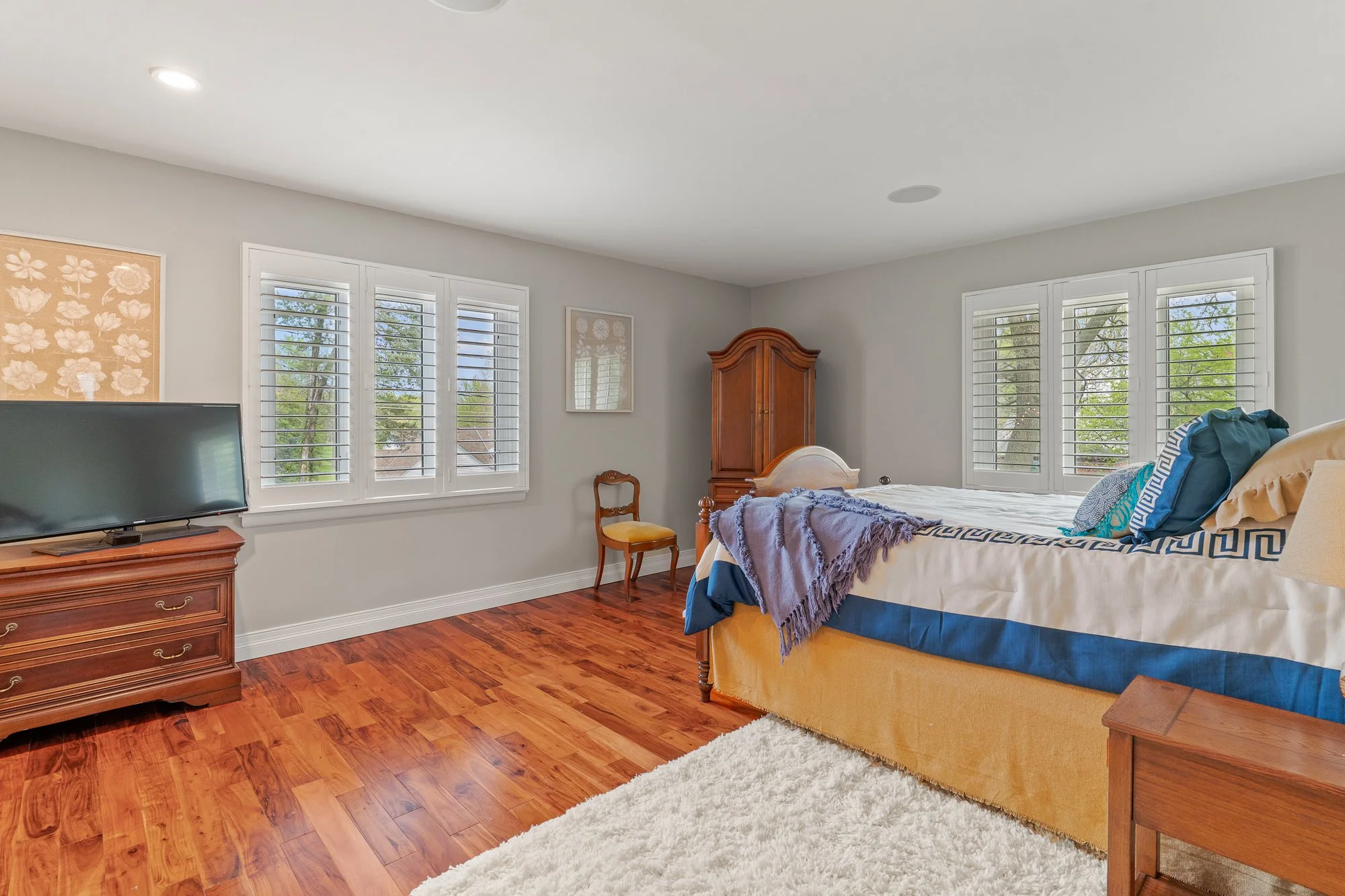 A bedroom with a bed, wooden dresser, a TV on a wooden stand, and windows with white shutters, wooden flooring, and decorated with framed art.