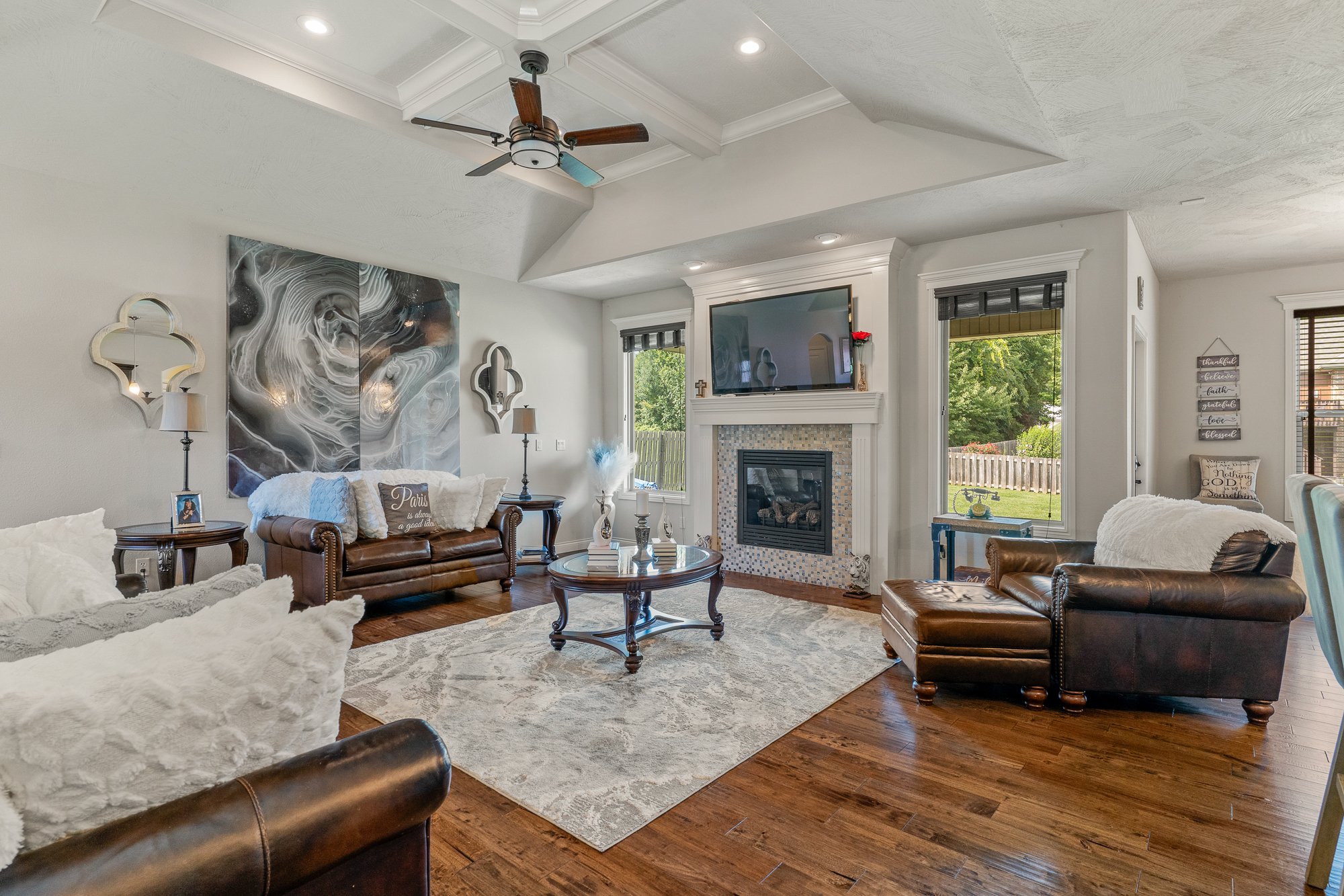 Living room with white walls, hardwood floors, leather sofas, a fireplace with a TV above, windows showing greenery outside, and modern decor including a large abstract wall art, mirrors, and a vase with red roses.