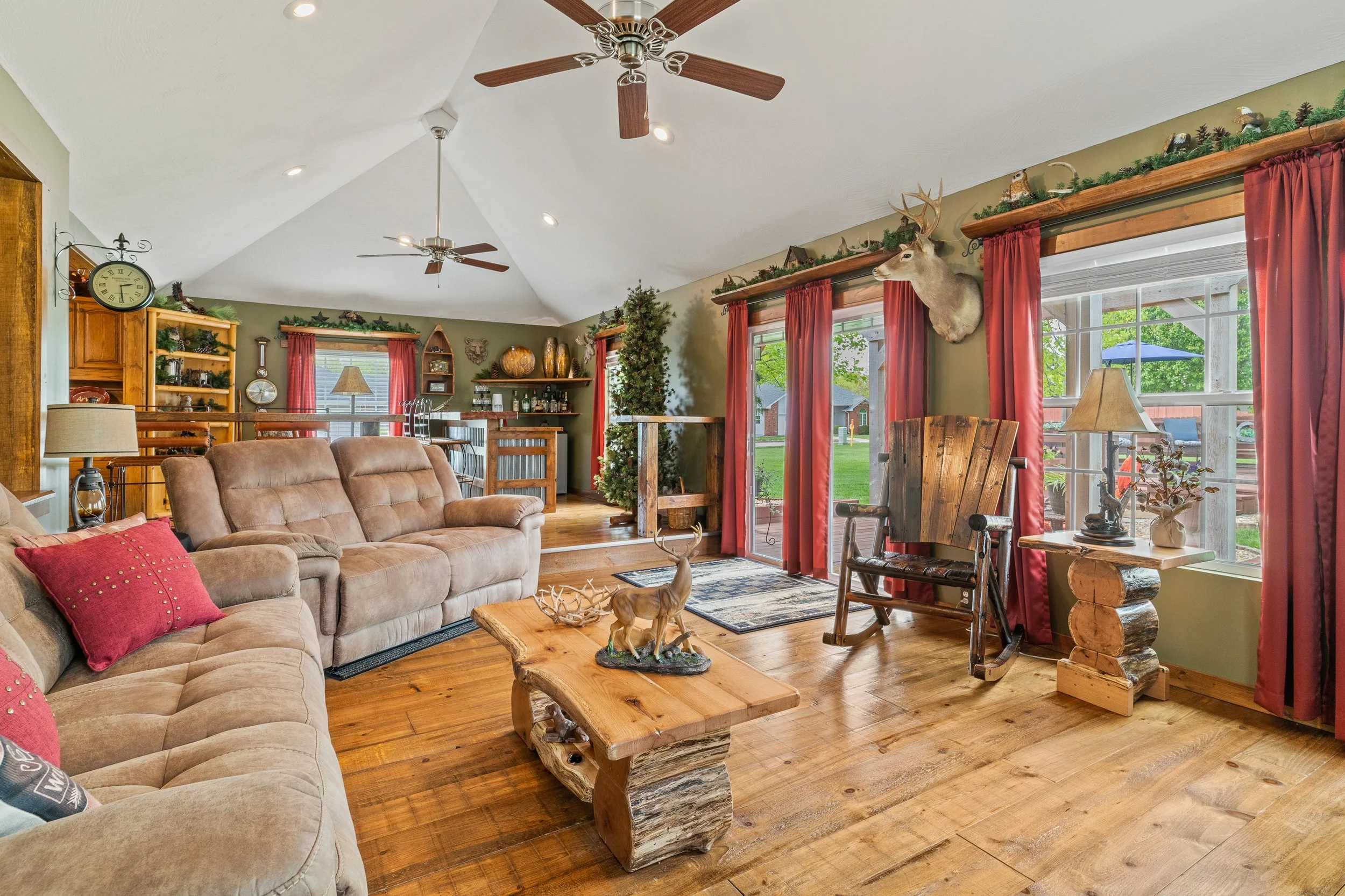 Living room with wooden floors, beige sofas with red pillows, large windows with red curtains, wooden coffee table with deer figurines, Christmas decorations, and a mounted deer head on the wall, with a view of a porch outside.