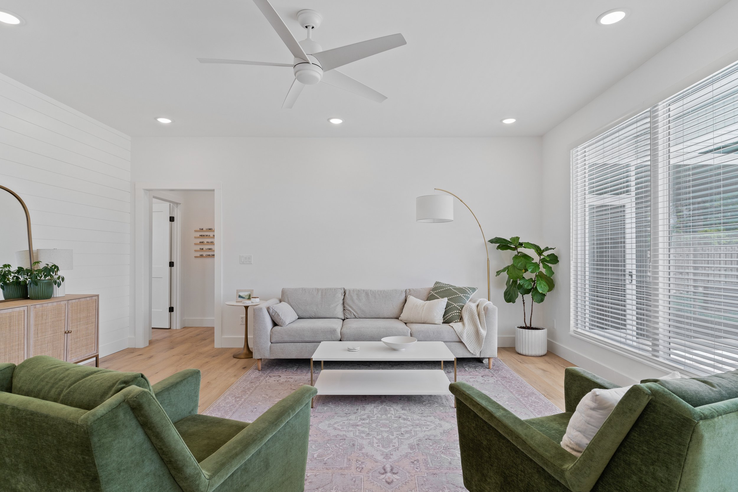 Living room with white walls, a gray sofa, green velvet armchairs, a white coffee table, large windows with blinds, natural light, a tall plant, and a ceiling fan.