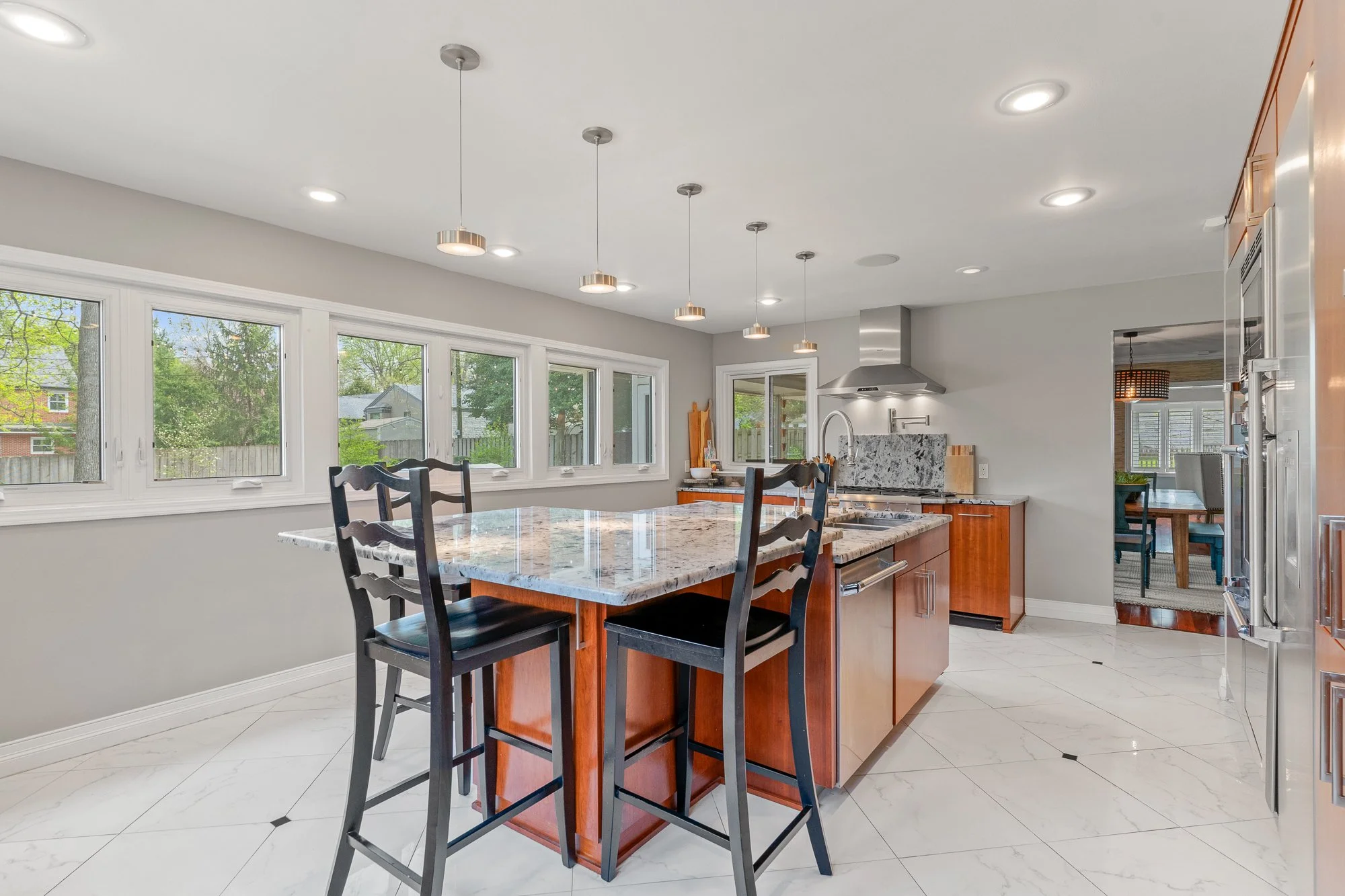 Modern kitchen with white tile floor, granite island counter, black chairs, large windows, stainless steel appliances, and a doorway leading to a dining area with a hanging lamp.