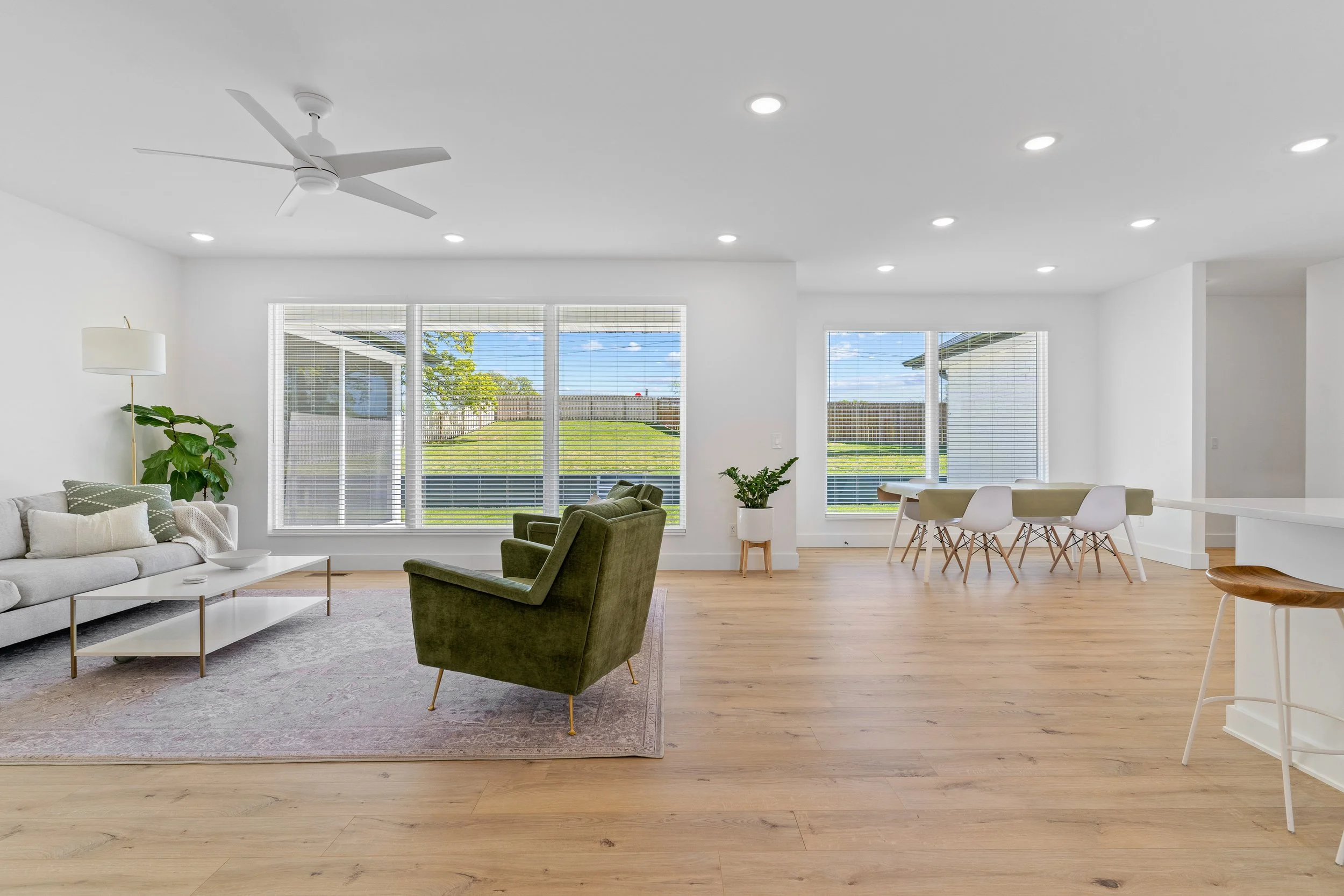 A spacious, open-concept living room and dining area with large windows showing a backyard with a lawn and blue sky. The room has a white ceiling with recessed lighting and a ceiling fan. Decor includes a light gray sofa, green armchair, white coffee