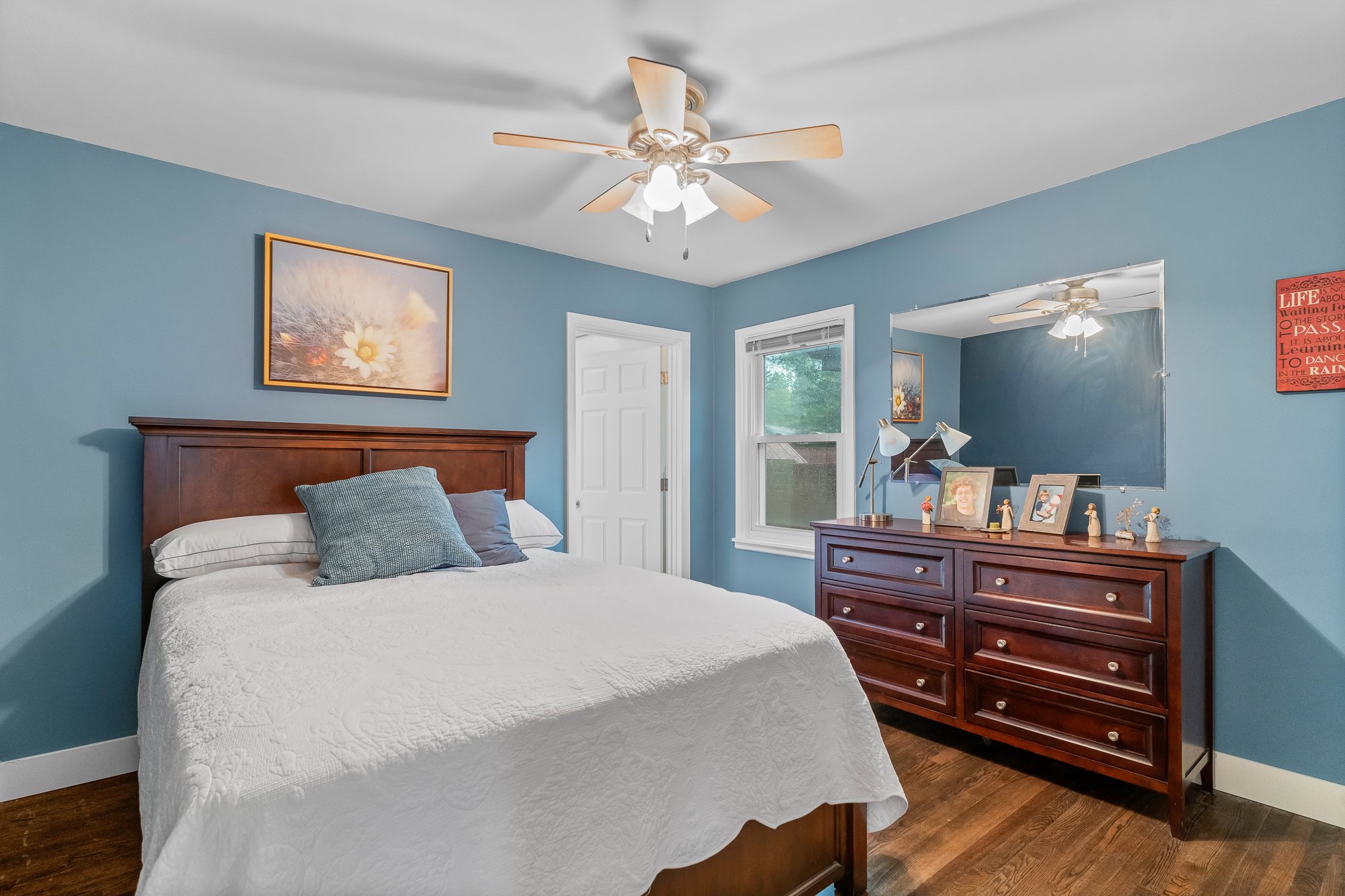 A bedroom with blue walls, a white ceiling fan, a bed with white quilt and pillows, a wooden dresser with framed photos, a window, and artwork on the walls.