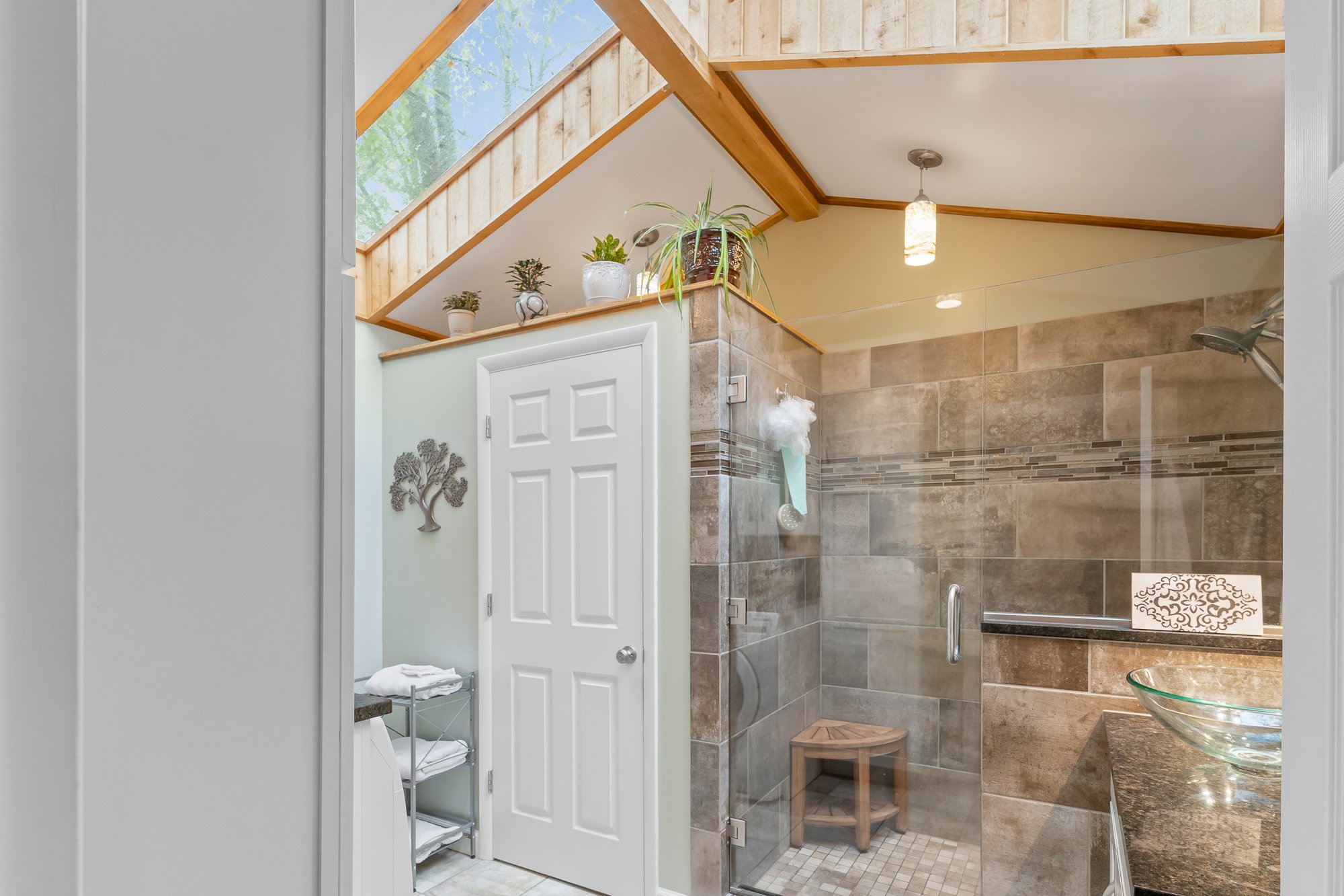 Bathroom with walk-in shower featuring brown and beige tile walls, a glass door, and a shower head. The ceiling has wooden beams and a skylight, with a small plant on the ledge. To the left, there is a white door and a decorative metal tree wall hang