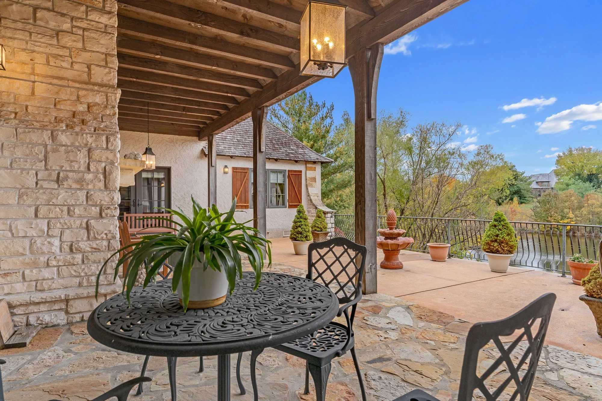 A covered outdoor patio area with wrought iron table and chairs, potted plants, and a decorative fountain, overlooking a scenic landscape with trees and a house in the distance under a bright blue sky.