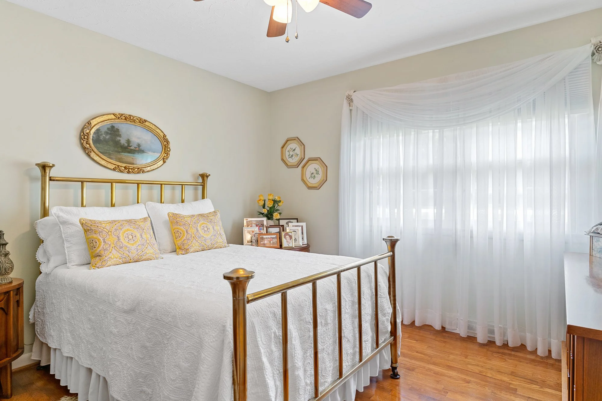 A bedroom with a brass bed, white quilt, yellow patterned pillows, and framed photographs on a bedside table. Above the bed are three picture frames and a landscape painting with a golden frame. The room has white walls, a ceiling fan, and a large wi