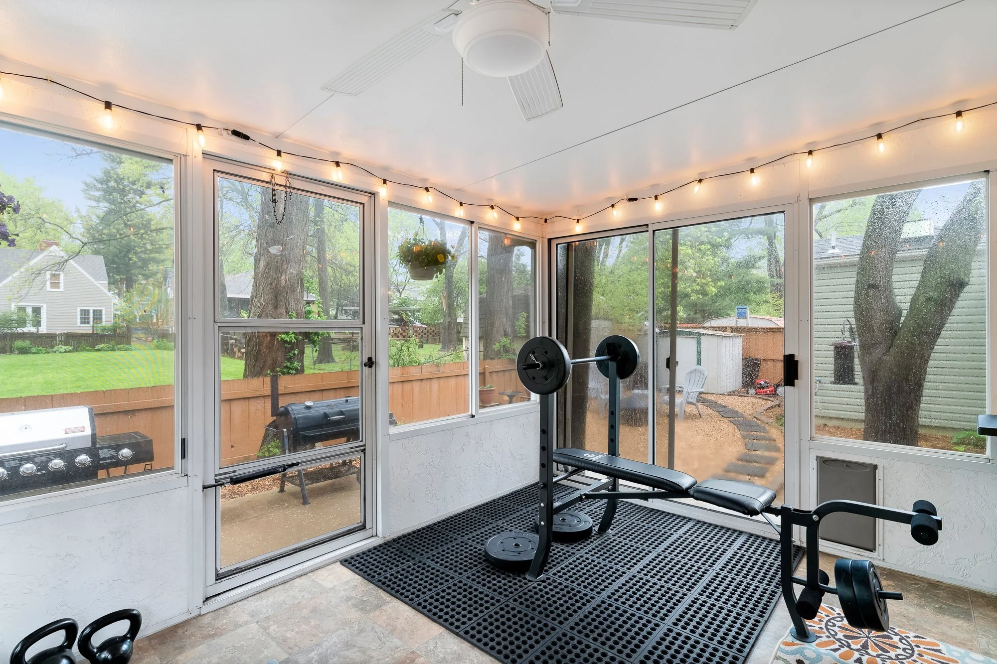 A screened-in porch with exercise equipment including a weight bench, barbell, and dumbbells. There is a black rubber mat and a small colorful rug. The porch has large windows and a ceiling fan. Outdoor view shows a backyard with a wooden fence, tree