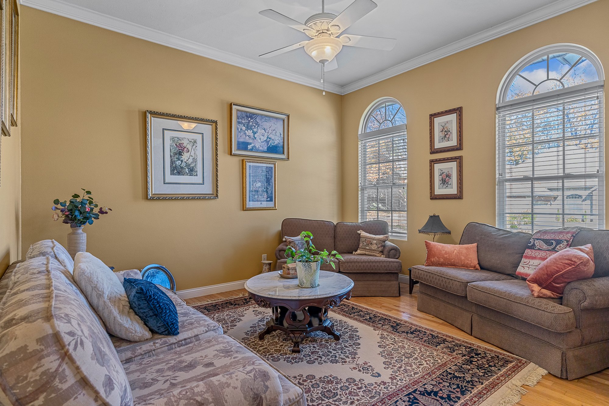 Living room with beige walls, large windows with blinds, a ceiling fan, and framed floral artwork. Furniture includes a beige sofa with pillows, a gray sofa, and a wooden coffee table with a potted plant.