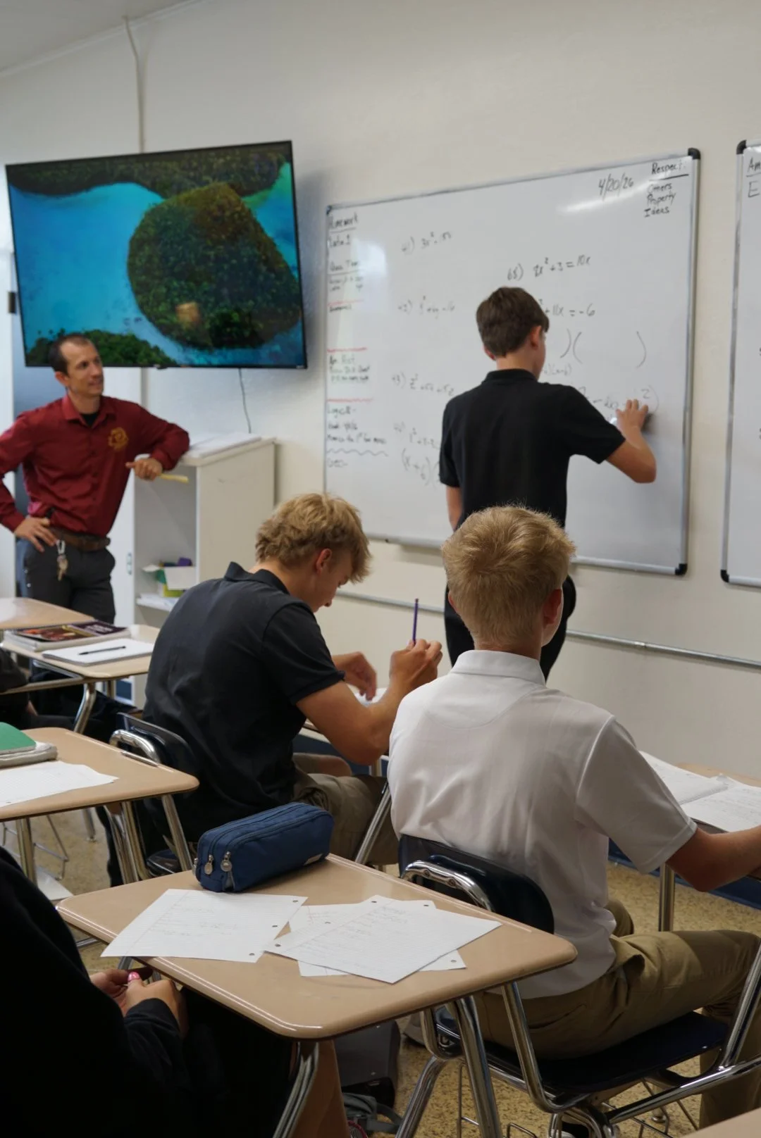 Students sitting at a table in a classroom, listening to a man standing by the window.
