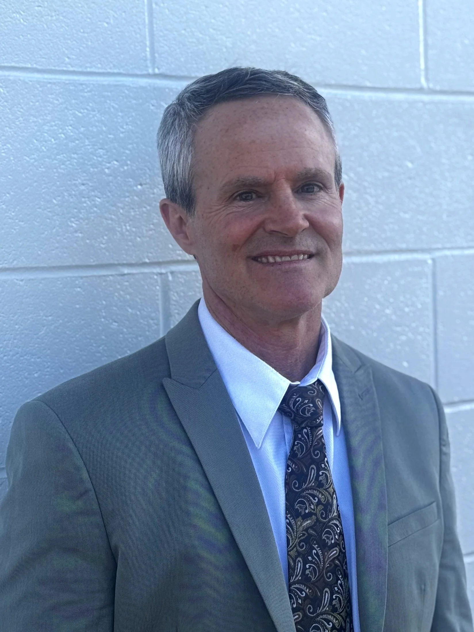 Man in a suit with a patterned tie standing in front of a white brick wall.