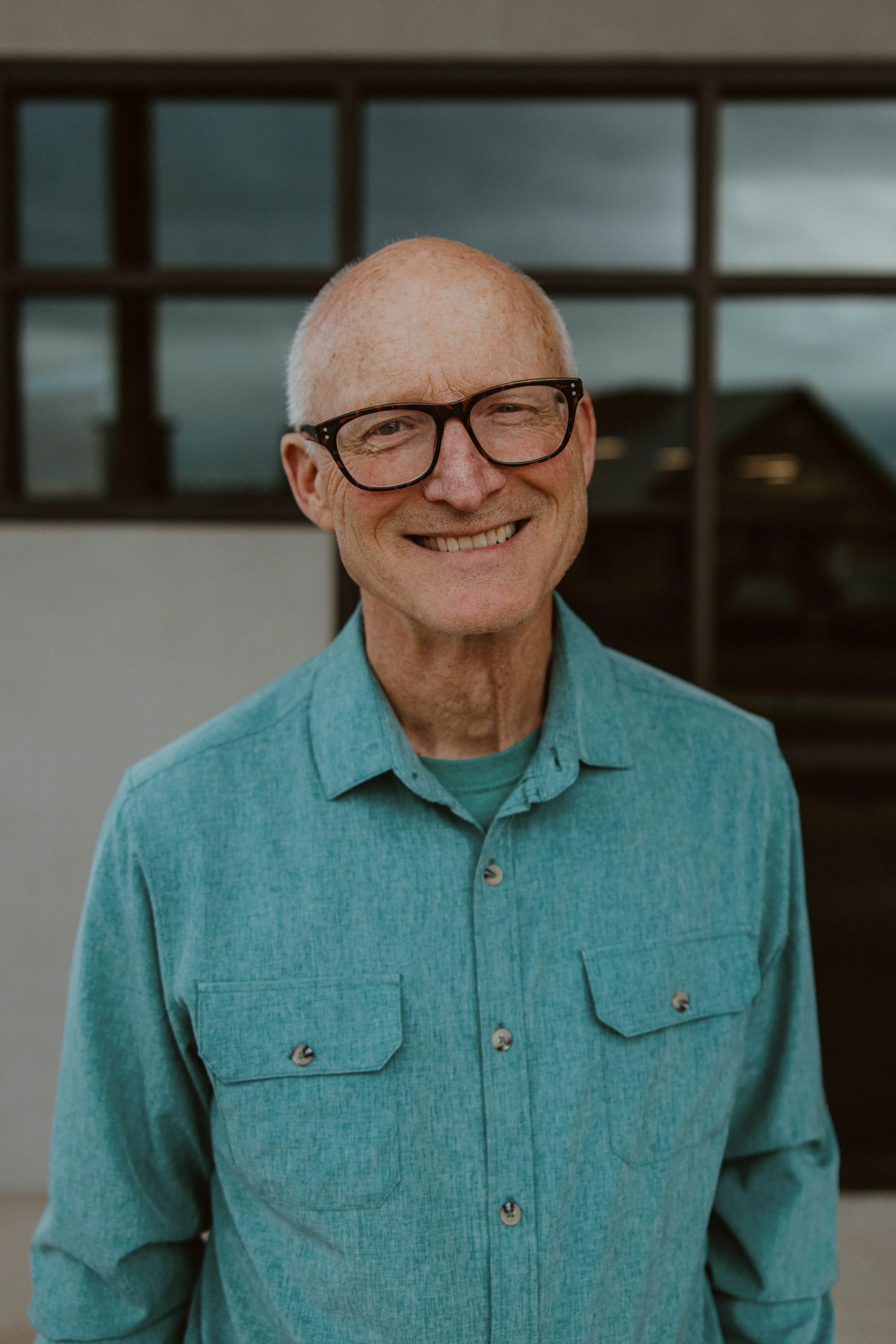 Smiling older man wearing glasses and a turquoise button-up shirt, standing in front of a window.