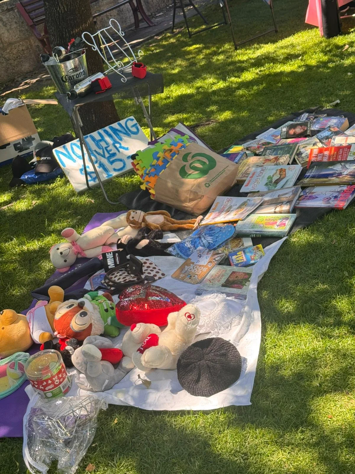 A yard sale setup on grass with toys, books, and household items displayed on a sheet and tables, with a sign that says "Nothing Over $1.00".
