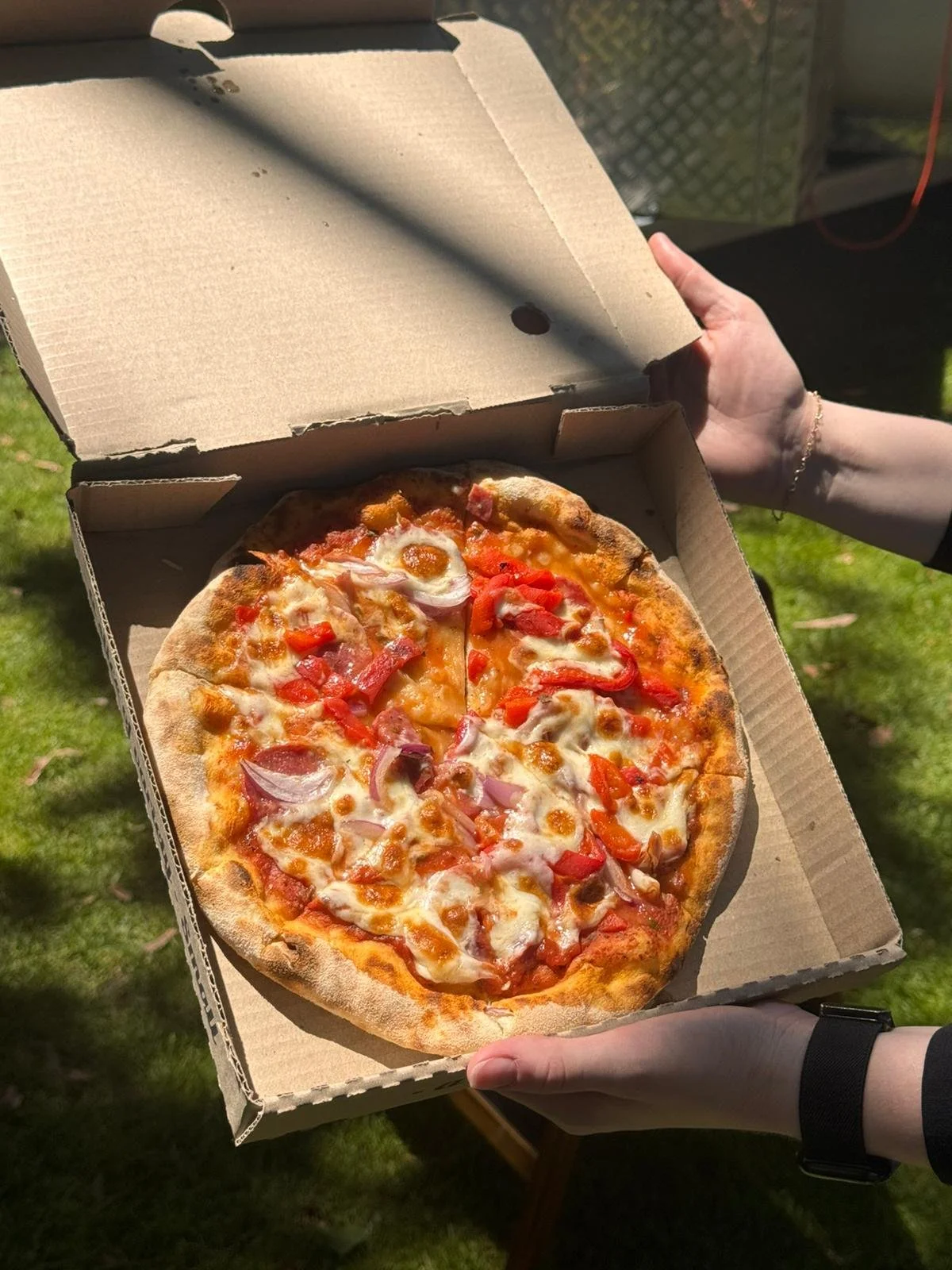 A pizza with cheese, red bell peppers, and onions in an open pizza box held by two people outside.