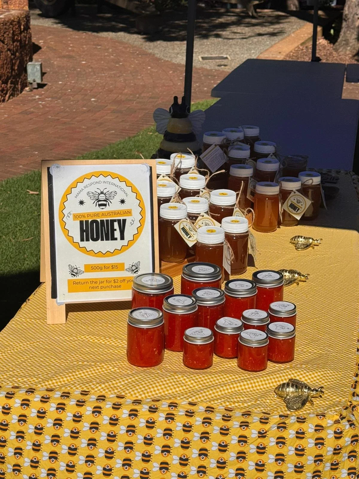 A display of small jars of honey for sale, with a sign indicating 100% pure Australian honey, 500 grams for $15, at an outdoor market table decorated with bee-themed fabric.