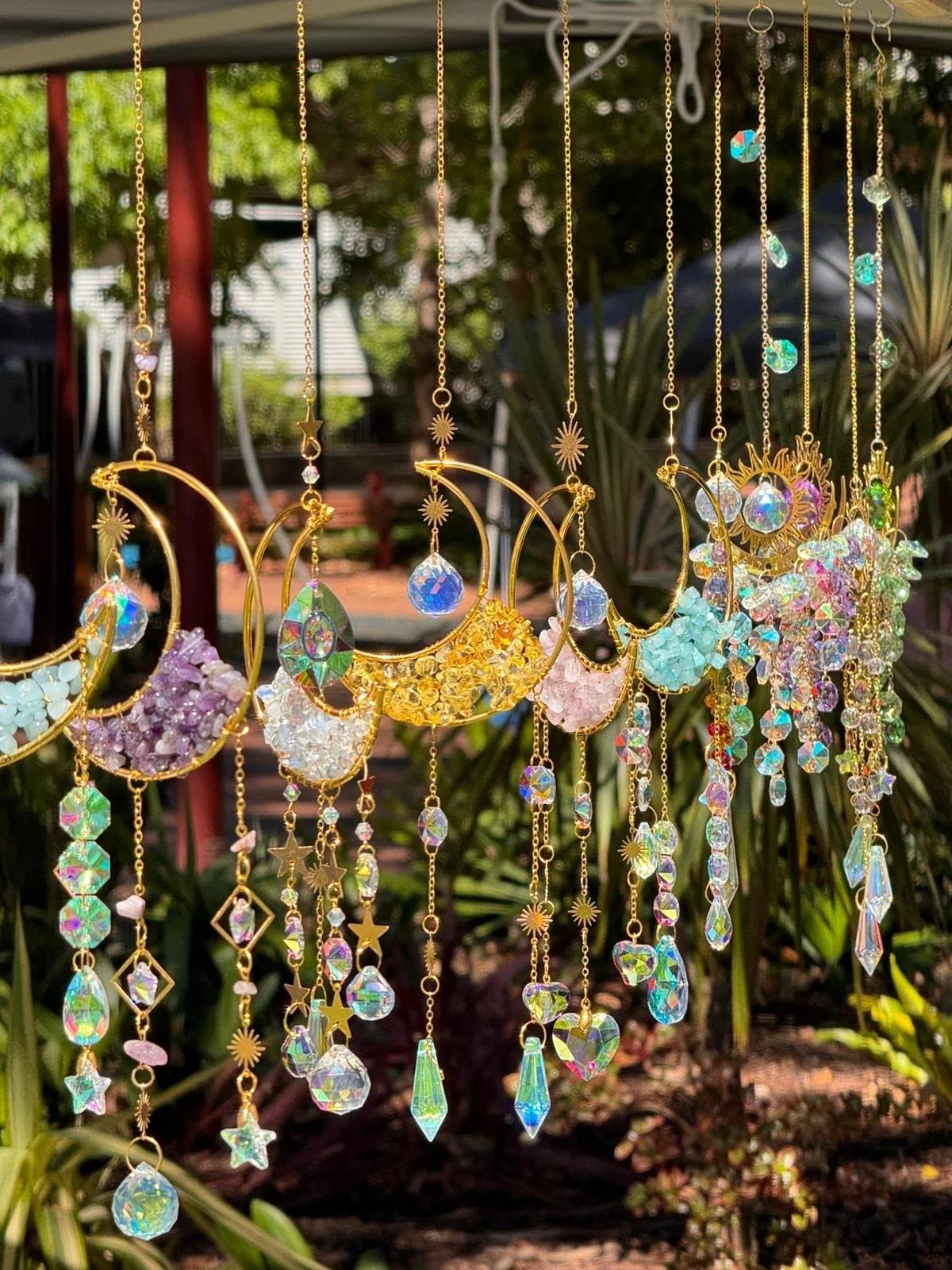 A display of decorative necklaces with gold chains, crescent moon shapes, and various colorful glass or crystal pendants hanging outdoors.