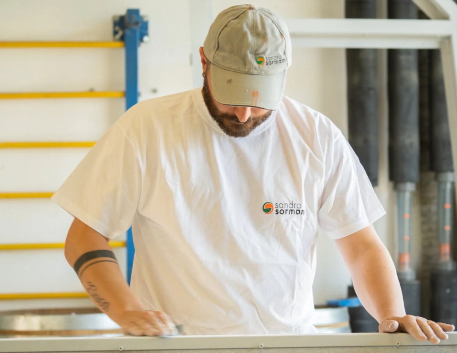 Un uomo che lavora in una bottega, indossa una tuta bianca con il logo di Sandro Sormani, una cappellino e si sta occupando di un'attività artigianale presso un banco di lavoro.