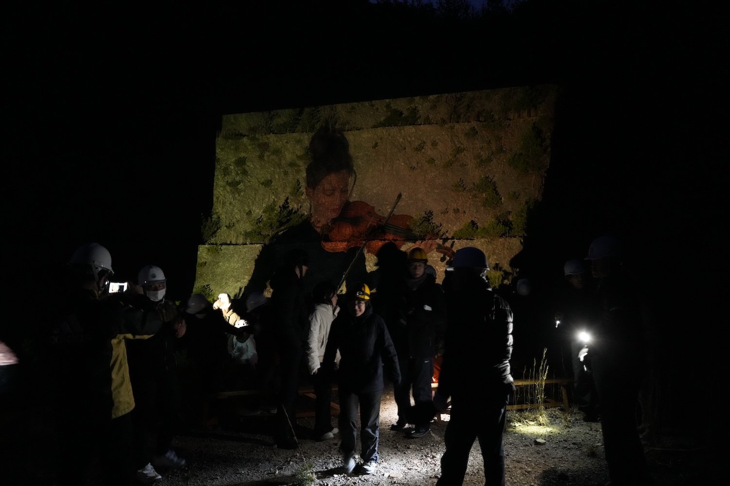 A projected violin performance illuminates the rock face of the former Shiroyama quarry at night while visitors gather to watch. 夜の城山採石場跡地で、岩壁に投影されたヴァイオリン演奏を集まった人々が鑑賞する様子。