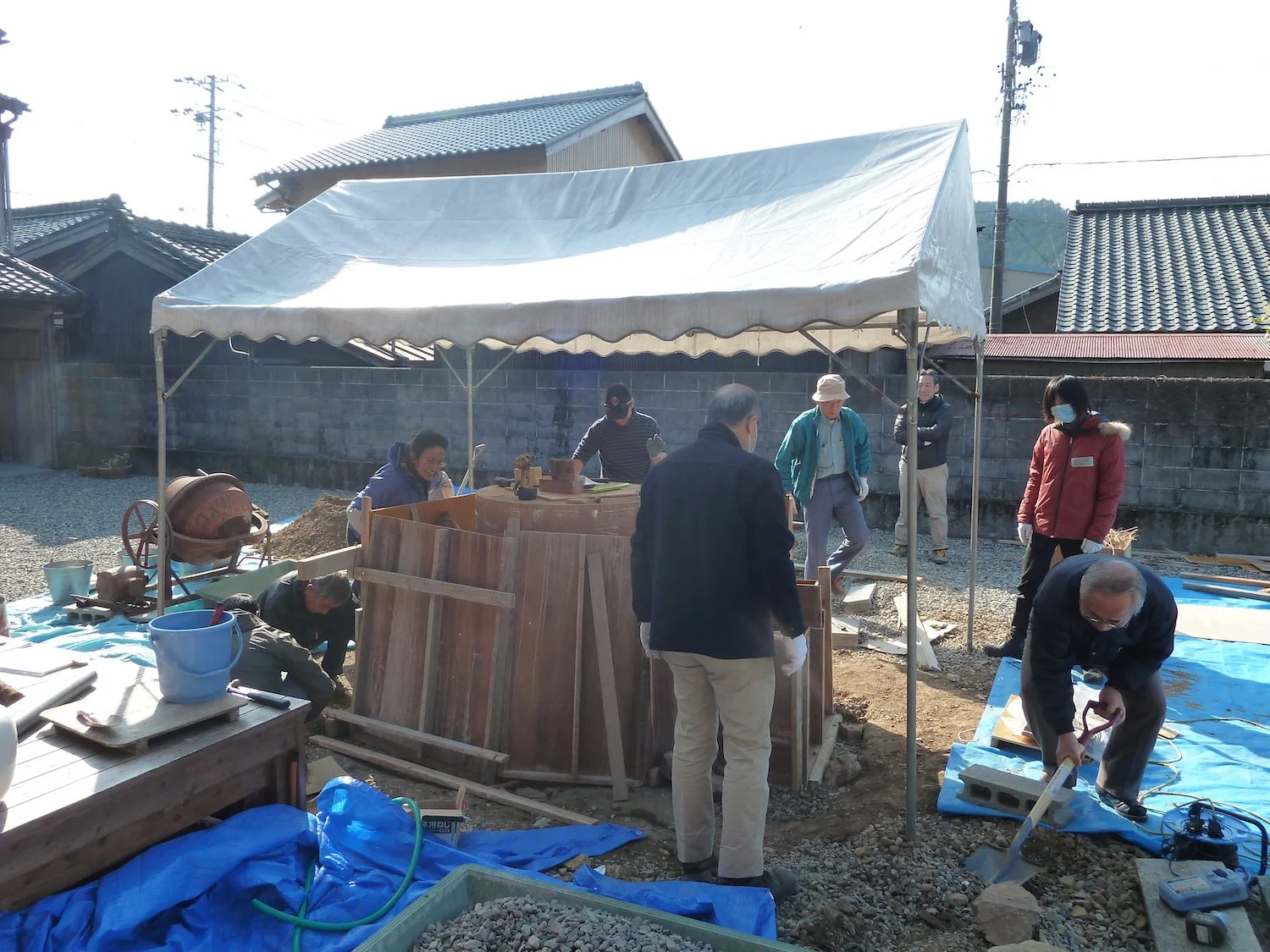 Participants work around wooden formwork beneath a temporary canopy during the construction of CAMINO in Kiinagashima. 紀伊長島での《CAMINO》制作中、仮設テントの下で参加者が木製の型枠を囲みながら共同作業をしている様子。