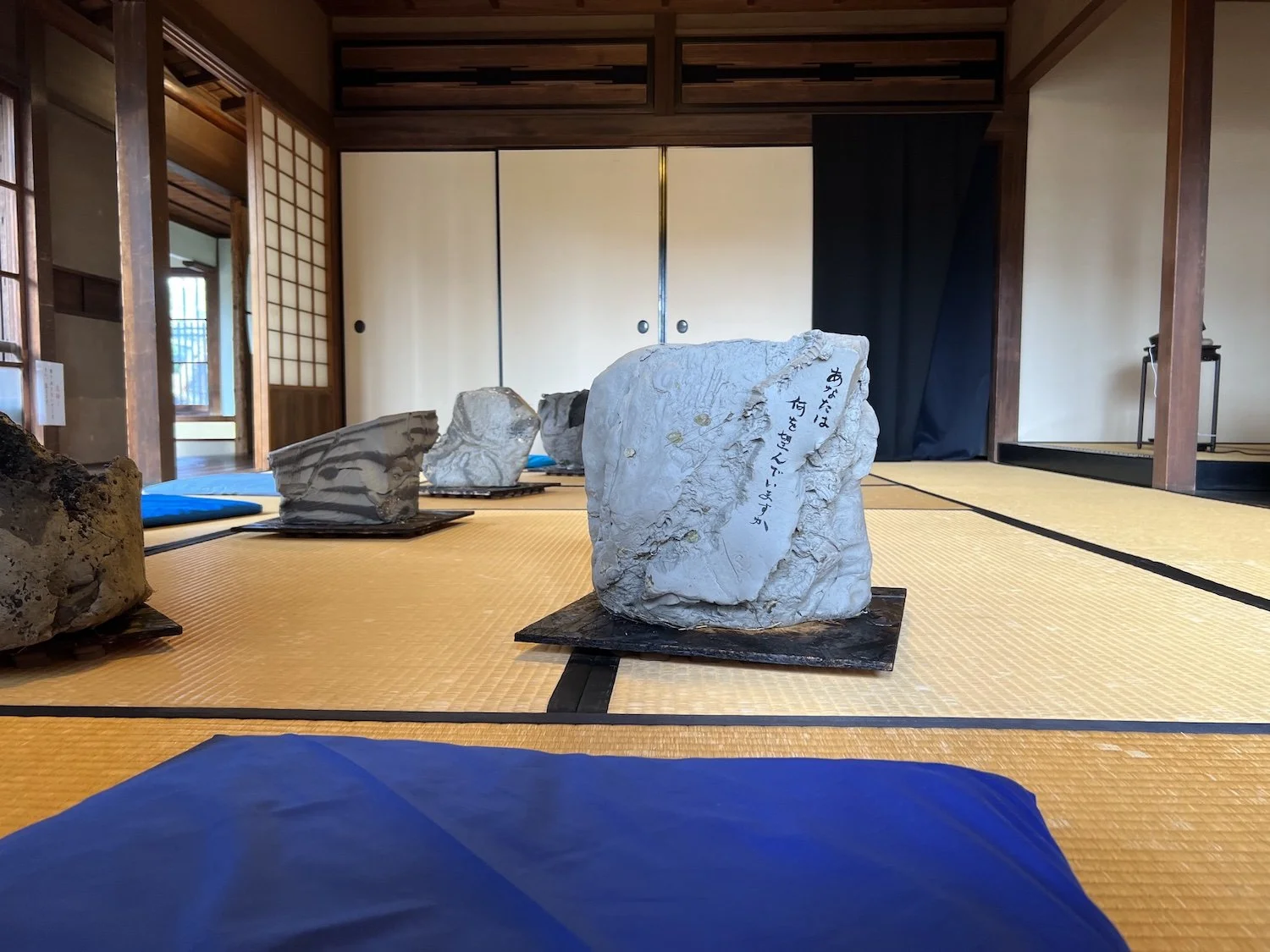 Several raw-clay sculptural forms arranged across tatami mats in the Sealed by Time installation at the Former Nakano Hanroku Residence, with blue fabric in the foreground. / 旧中埜半六邸の《Sealed by Time》で、畳の上に配置された複数の生の粘土による造形と、手前に広がる青い布。