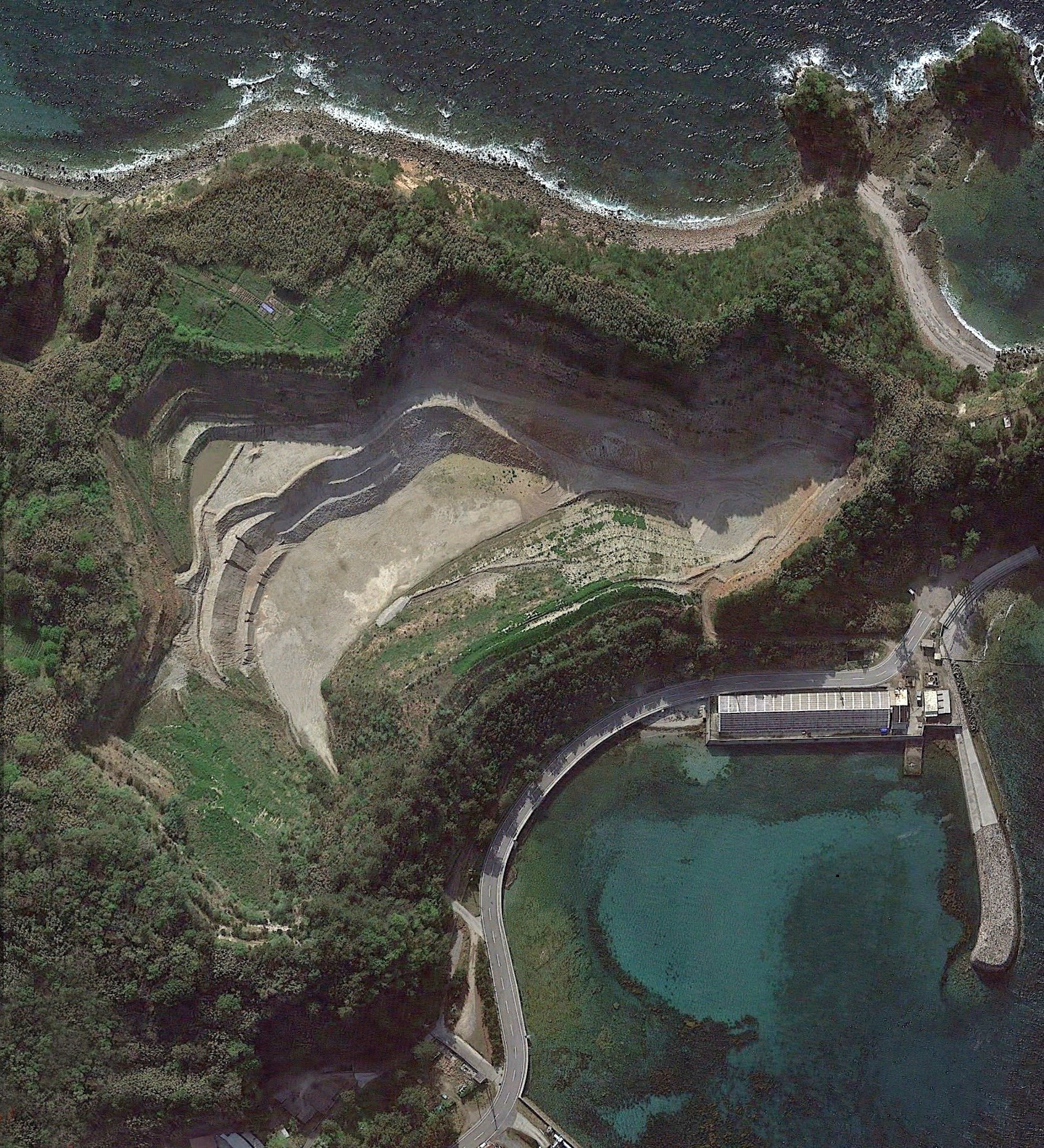 Aerial Google Earth view of the former Shiroyama quarry site on Himeshima Island, showing exposed rock terraces, surrounding forest, and nearby sea. 姫島の旧城山採石場跡地をGoogle Earthで上空から見た画像。露出した岩の段丘、周囲の森、近くの海が見える。