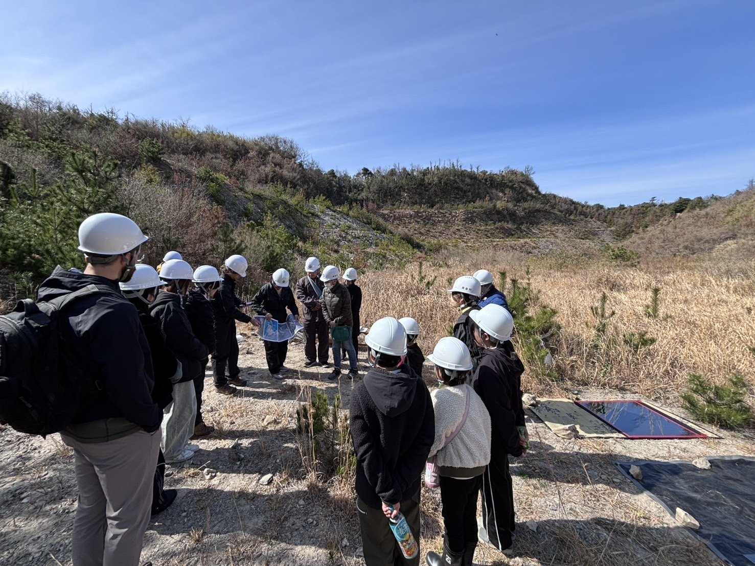 A group of people wearing helmets visits the former Shiroyama quarry site on Himeshima Island, observing the landscape as part of the ongoing Shiroyama Labyrinth dialogue. ヘルメットを着用した参加者が姫島の旧城山採石場跡地を見学し、城山ラビリンス構想に向けた継続的な対話の一環として風景を観察している様子。