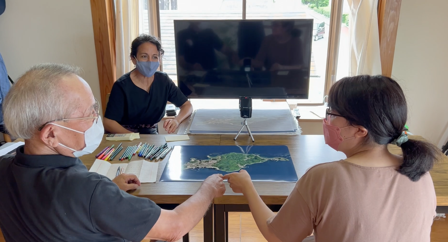 A grandfather and granddaughter discuss Himeshima around an island map, with a remote participant joining by screen. / 姫島の地図を囲んで語り合う祖父と孫娘。画面を通してオンライン参加者も加わっている。