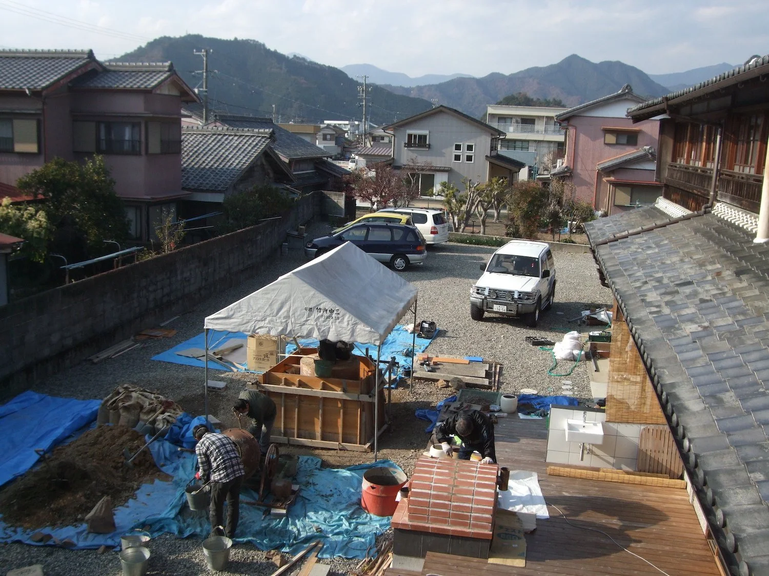 Elevated view of the CAMINO construction site at Yugakutei, showing a temporary white canopy, blue tarps, tools and materials in the courtyard, with houses and mountains beyond. ゆうがく邸での《CAMINO》制作現場を上方から見た様子。中庭に白い仮設テント、青いシート、工具や材料があり、奥に住宅と山並みが見える。