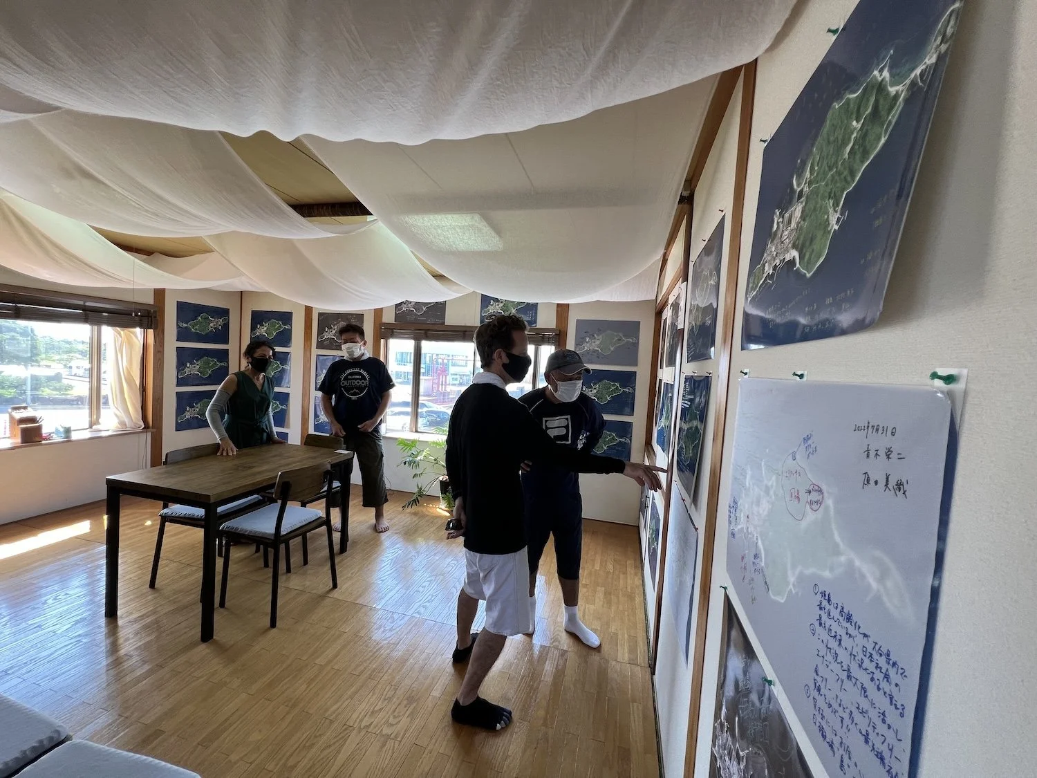 Visitors viewing maps and handwritten contributions displayed throughout the Island Atlas exhibition space in Himeshima. / 姫島での《島のアトラス》展示会場で、壁に展示された地図や手書きの記録を見る来場者たち。