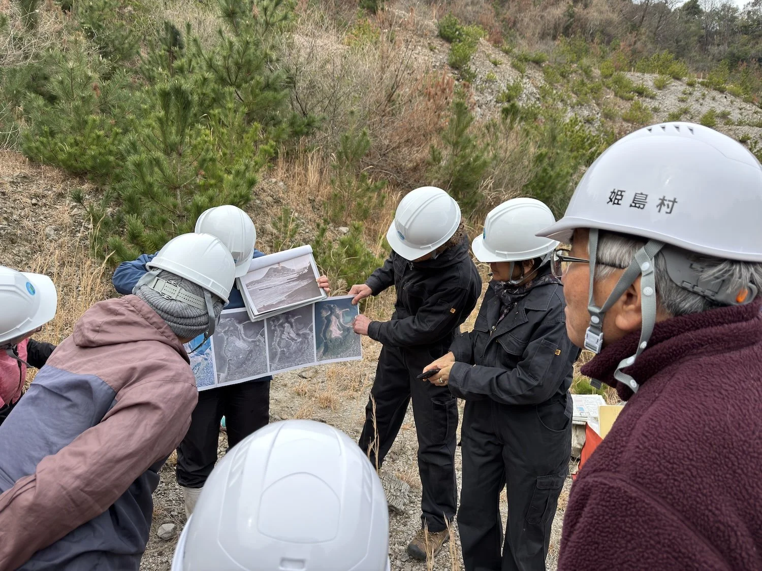 Participants wearing safety helmets examine geological information with a geologist at the former Shiroyama quarry in Himeshima. 姫島の城山採石場跡地で、ヘルメットを着用し、地質学者と地質資料を確認する参加者たち。