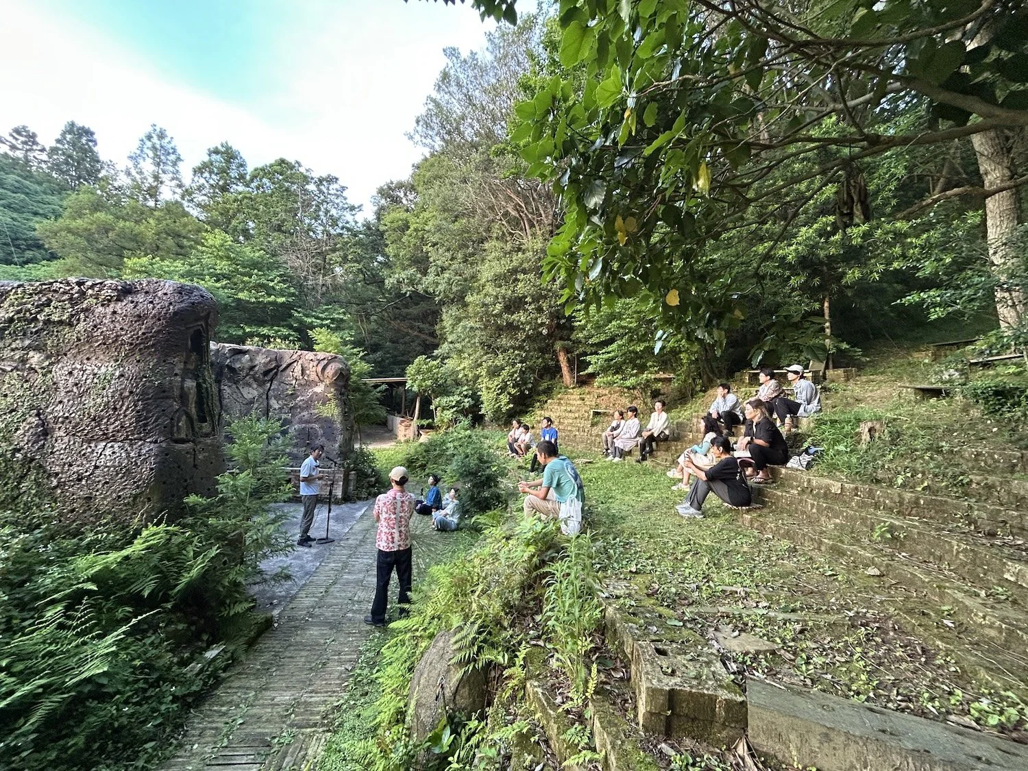 People sit on the terraced seating at Yama no Hiroba while listening to a speaker during a 360° Open Hiroba gathering. 山の広場®の段状の客席に座り、360°オープン広場で話を聞く参加者たち。