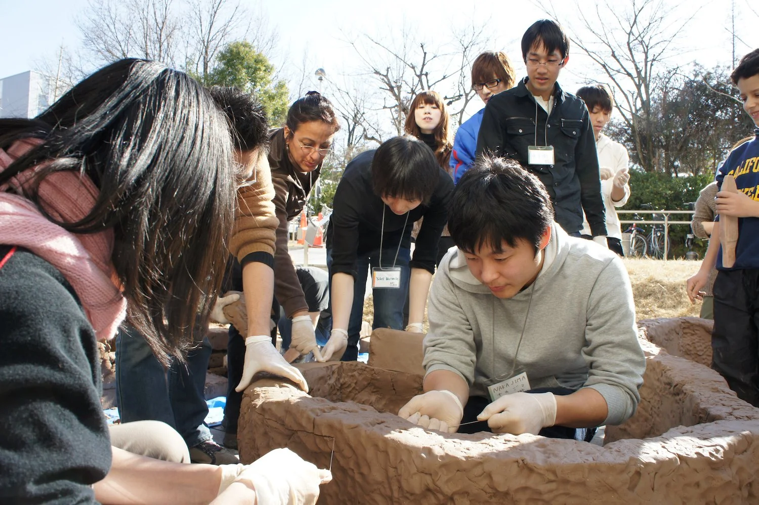 Participants work around the raw-clay structure during the Common Ground workshop beside the green Clas Gallery banner at Nagoya University. 名古屋大学の緑色のClas Galleryバナーのそばで、《Common Ground》の生土の構造体を囲み、参加者が共同制作している様子。