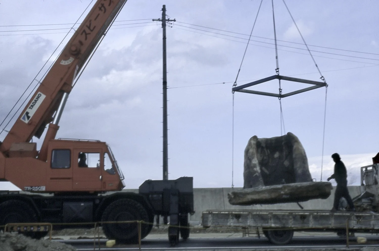 A crane lifting a large bear sculpture off a truck, with a person nearby during daytime under cloudy skies.